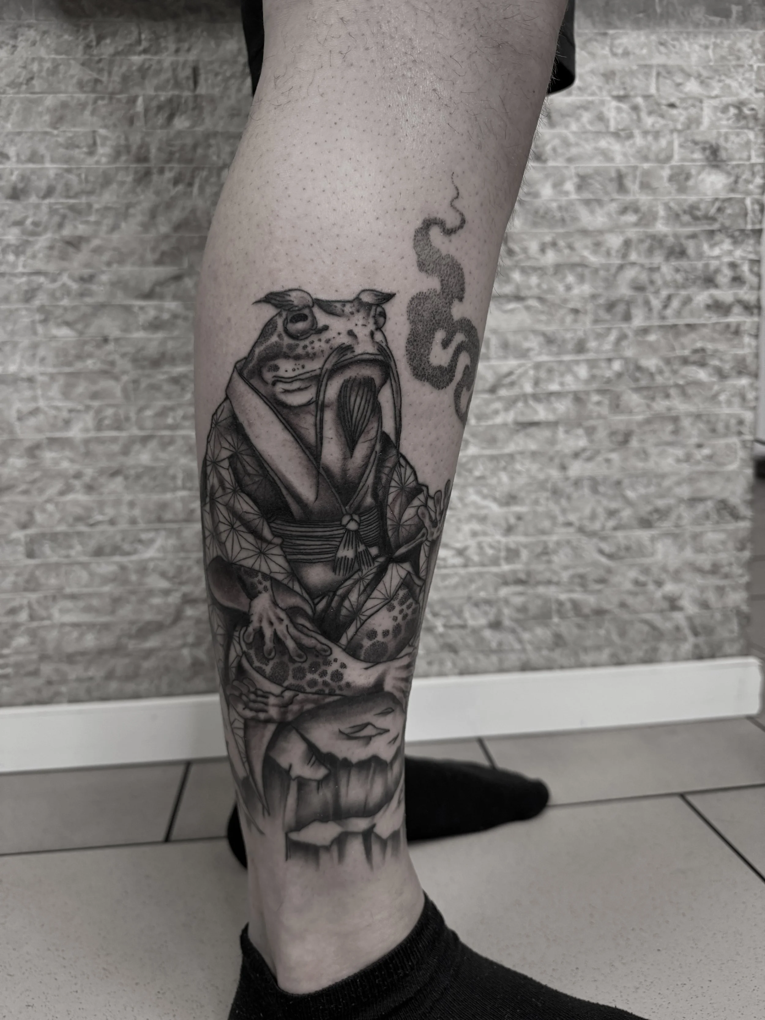 Schwarz-weiße Tattoo auf dem Unterschenkel einer Person, zeigt einen Frosch in traditionellem japanischem Stil mit einer Kimono, der auf einem Felsen sitzt, mit chinesischen oder japanischen Schriftzeichen neben ihm