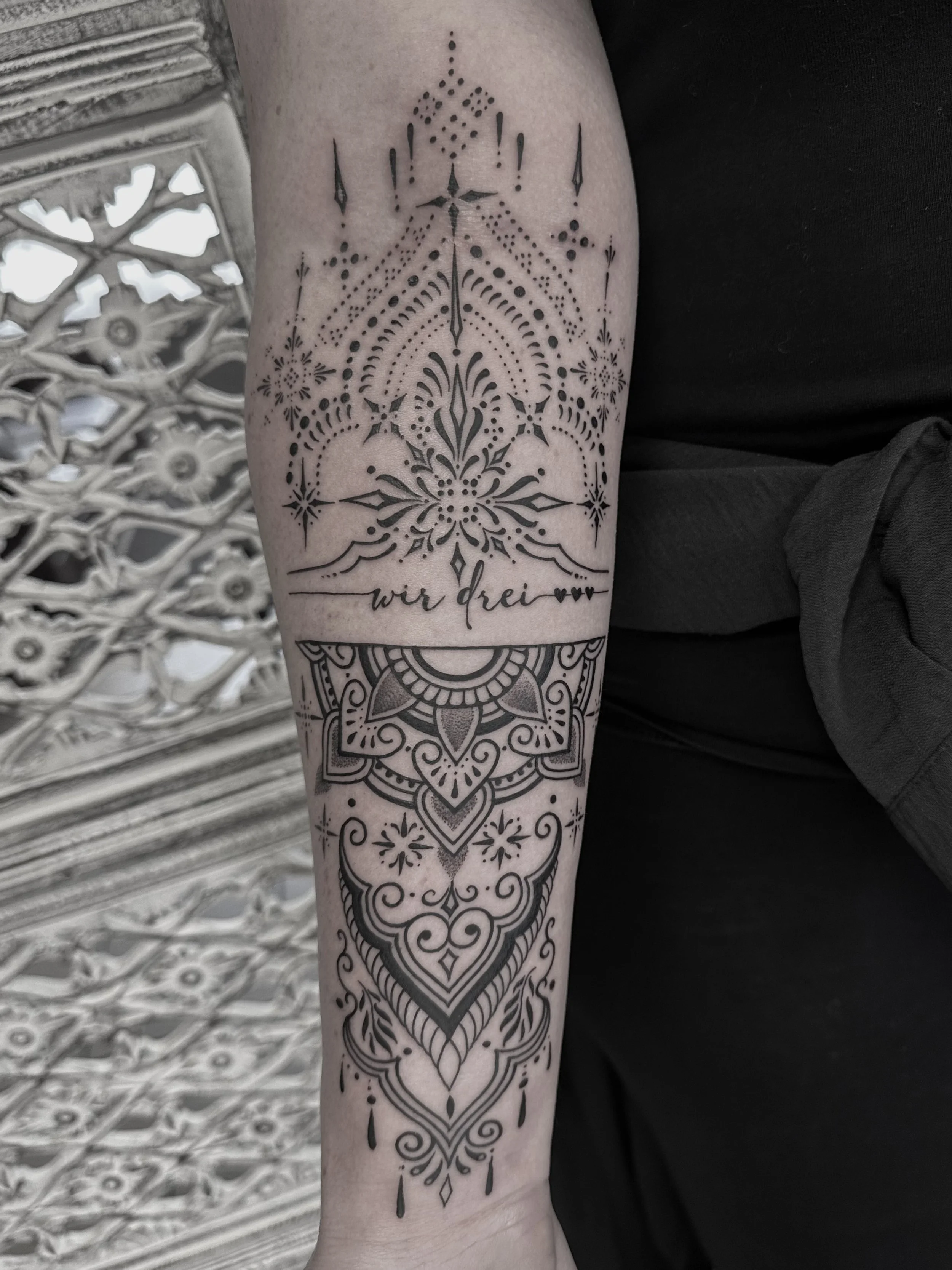 Schwarze Tattoo auf Unterarm mit geometrischen und floralem Design, darunter die Worte ‚wir drei‘ mit zwei kleinen Herzen.