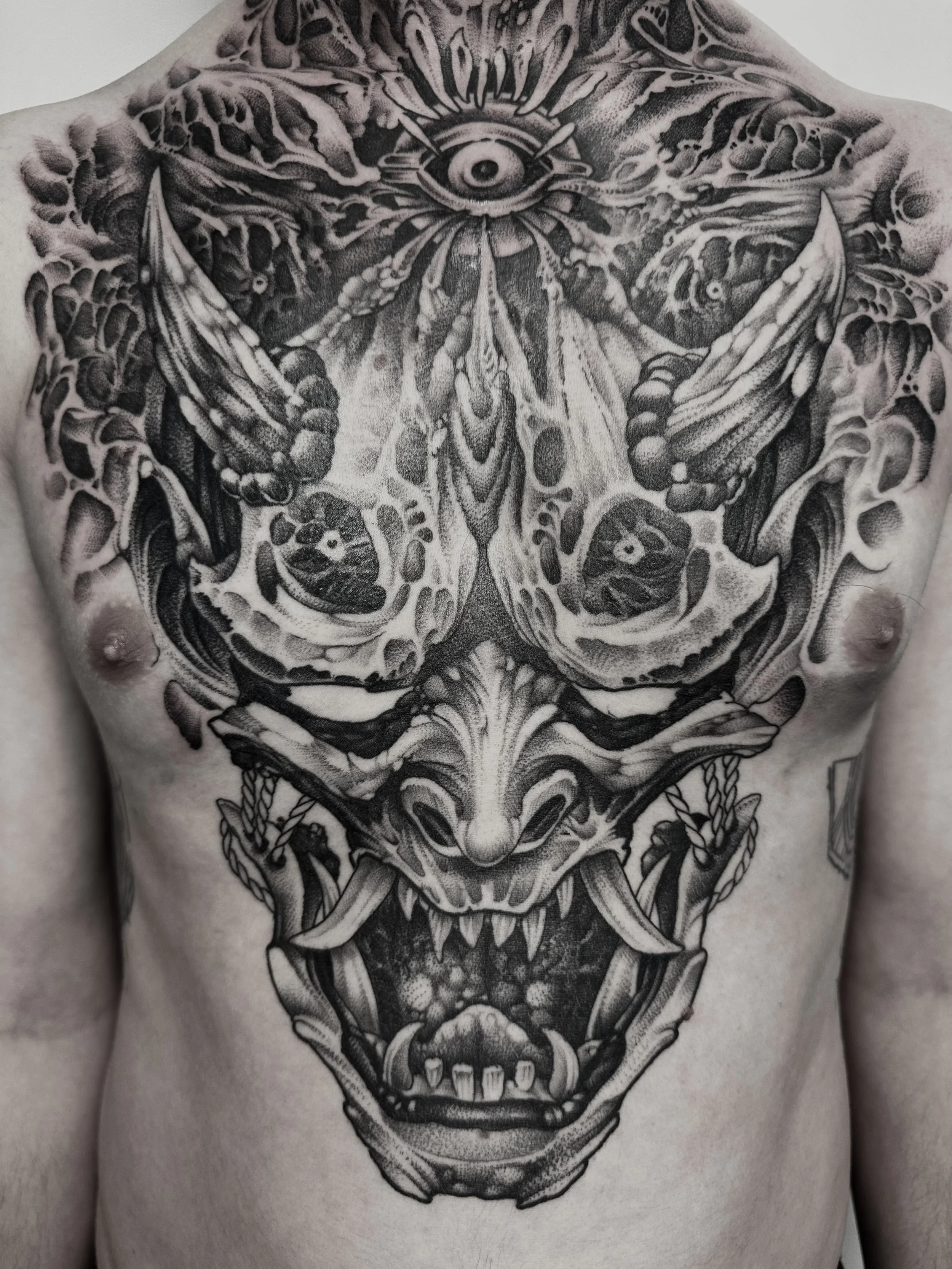 Schwarze Tätowierung eines dämonischen Gesichts auf der Brust, mit einem einzelnen Auge auf der Stirn und einem offenen Maul mit Zähnen.
