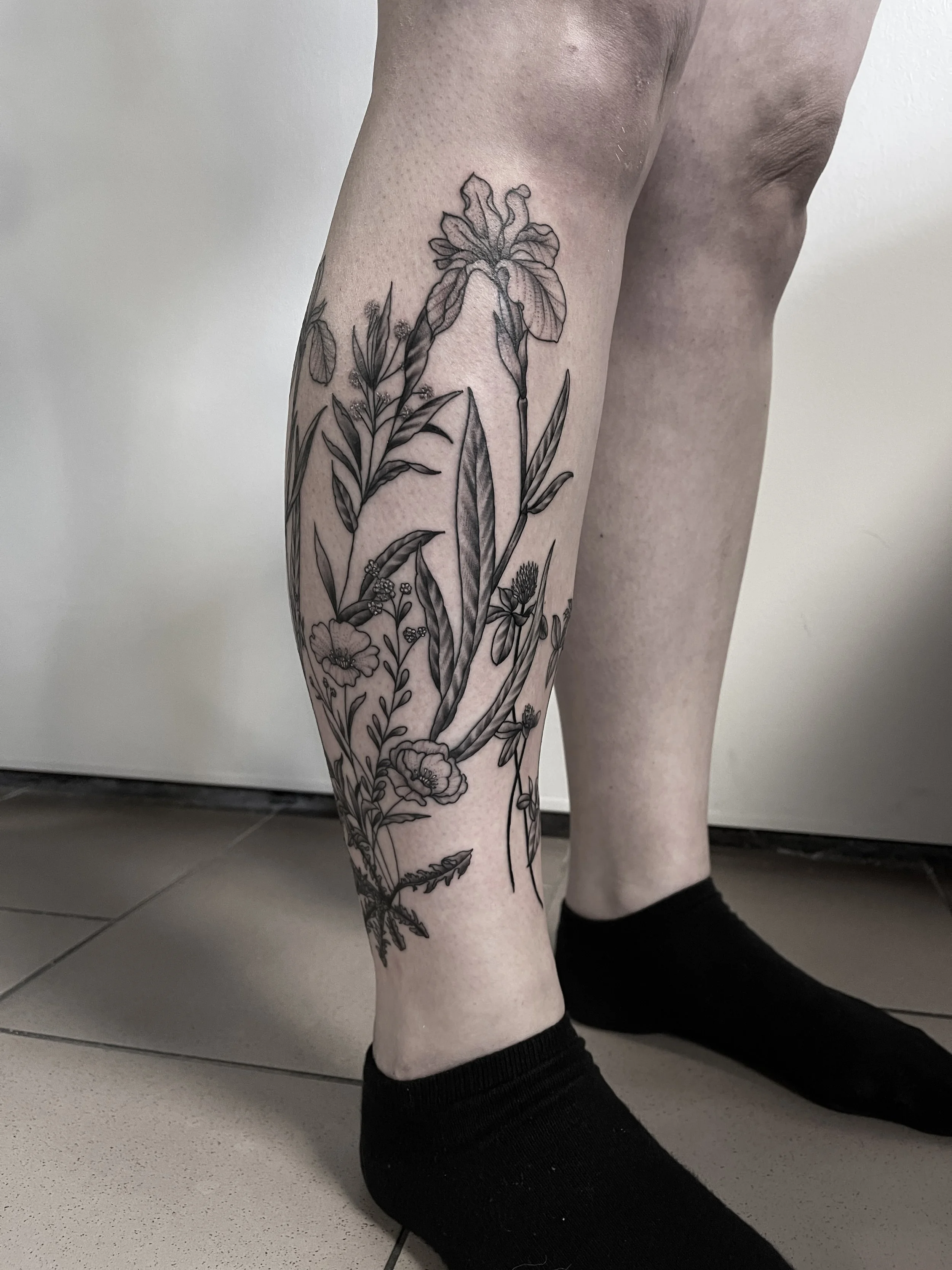 Schwarze Tätowierung eines Blumenmusters auf dem Unterschenkel einer Person, die schwarze Socken trägt.