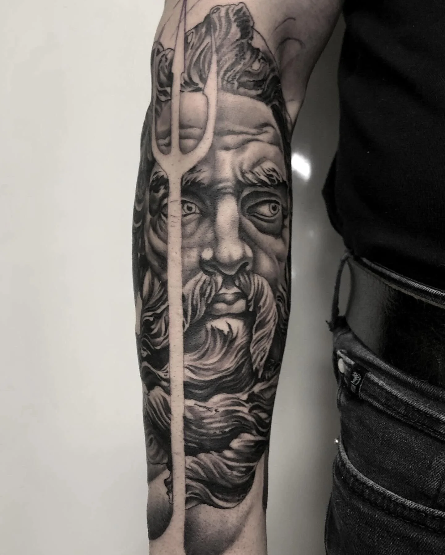 Schwarze Tattoo eines alten Mannes mit langem Bart und einer Wal, mit einer Statue und einem Dreizack im Hintergrund.