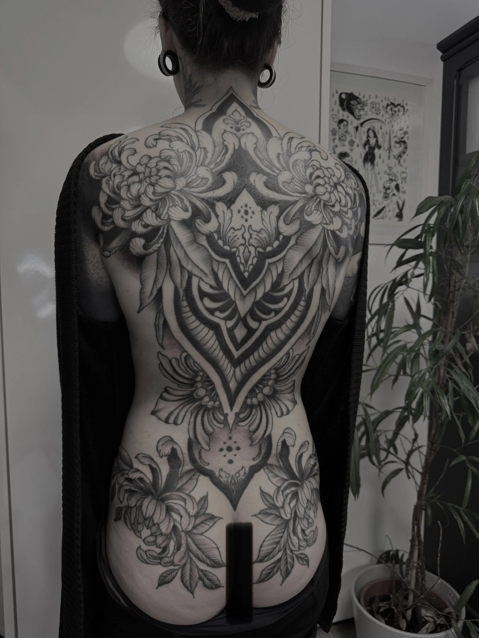 Rückenansicht einer Person mit umfangreichem schwarzen Thai-Tattoo mit Pflanzenmotiven auf Rücken, Schultern und unteren Rücken, in Innenraum mit Pflanzen und Wandkunst