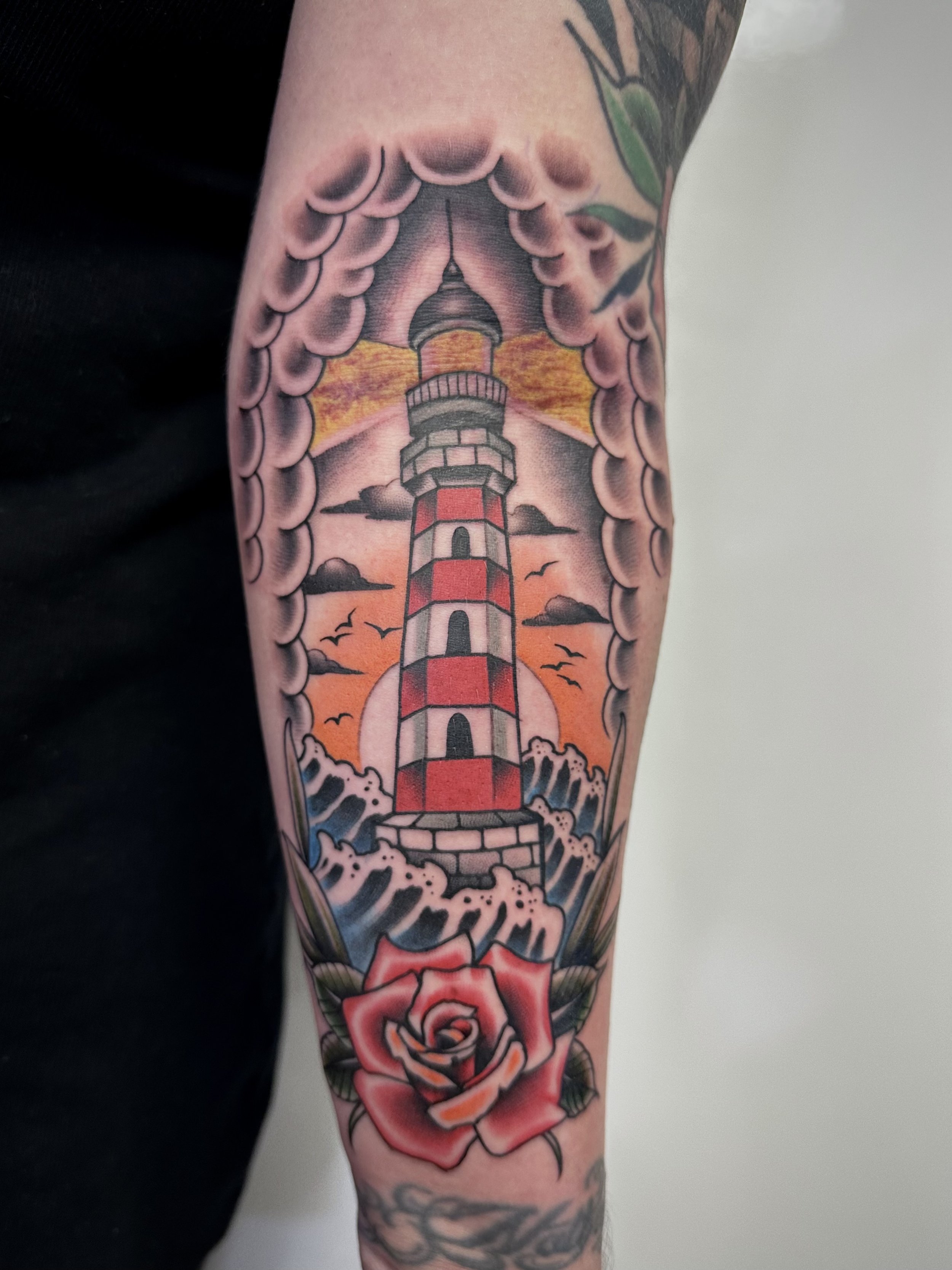 Tattoo eines Leuchtturms in der Mitte mit Wolken, Vögeln und einem Sonnenuntergang im Hintergrund, darunter eine rosafarbene Rose mit grünen Blättern.