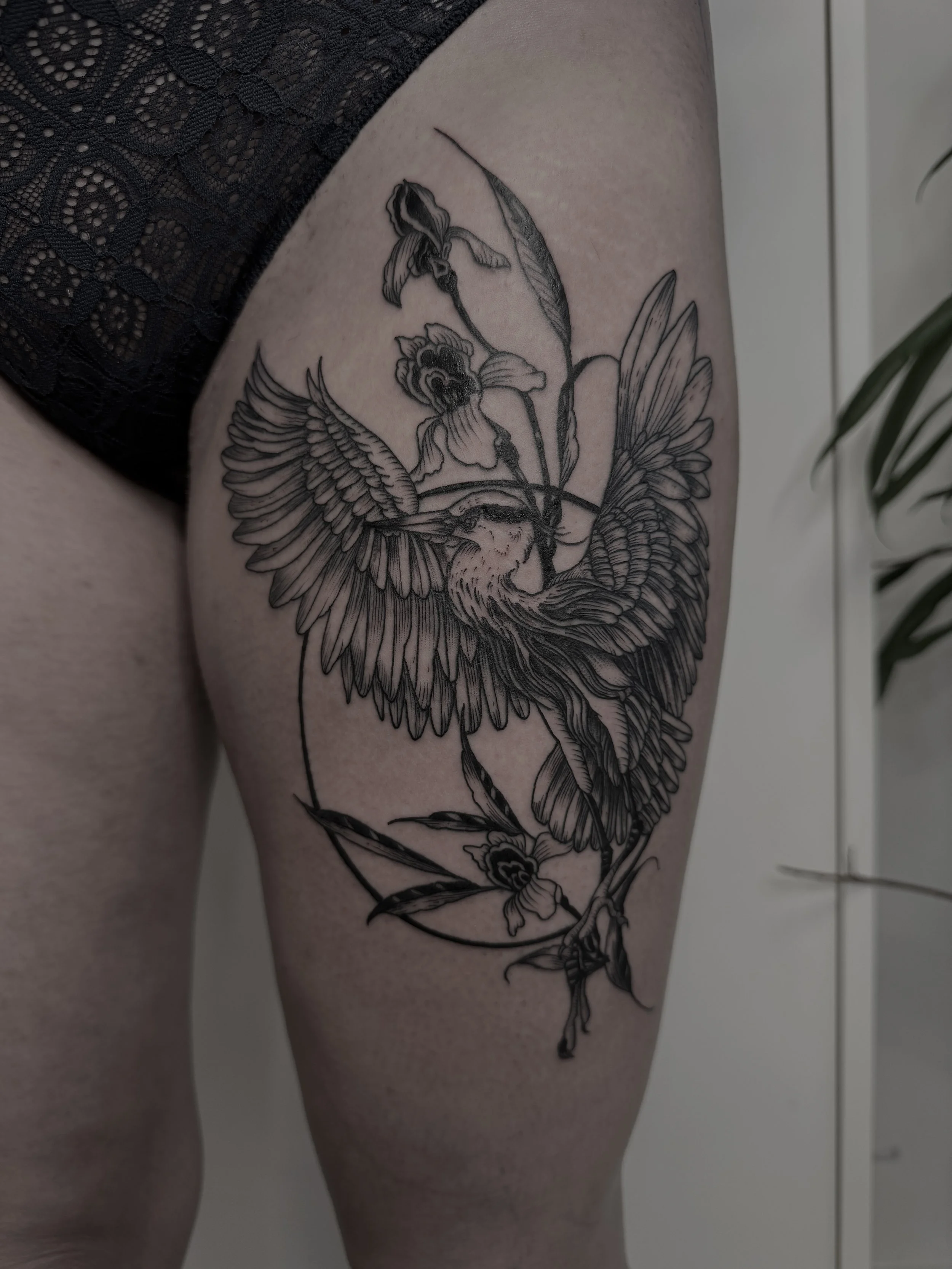 Detaillierte schwarze Tätowierung eines Vogels, umgeben von Blumen, auf dem Oberschenkel einer Person.