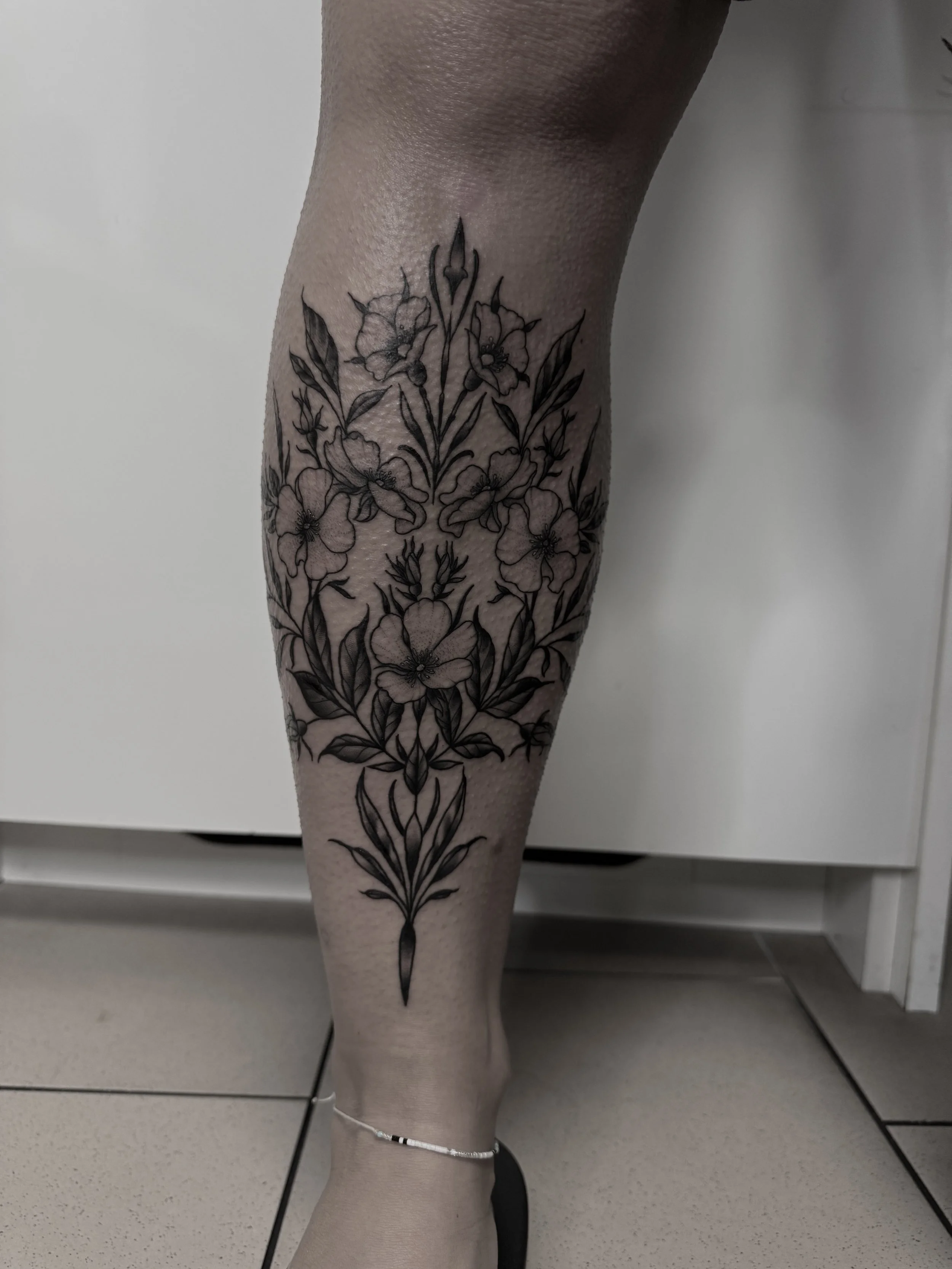 Schwarz-weiße Tätowierung eines Blumenmusters auf dem Unterschenkel eines Menschen, mit mehreren verschiedenen Blüten und Blättern.