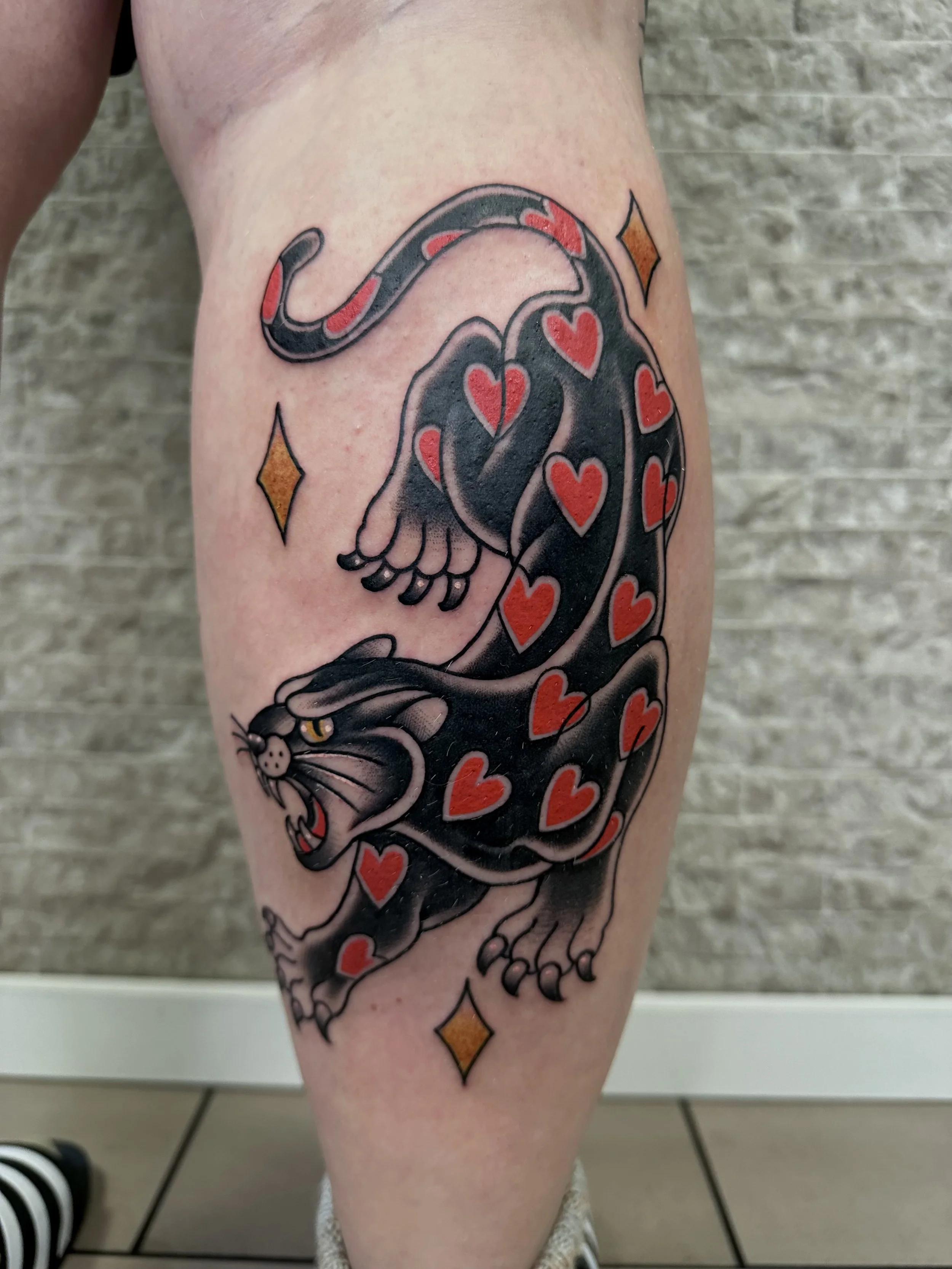 Tattoo eines schwarzer Panther mit roten Herzen auf dem Körper, umgeben von kleinen orangefarbenen Diamanten.