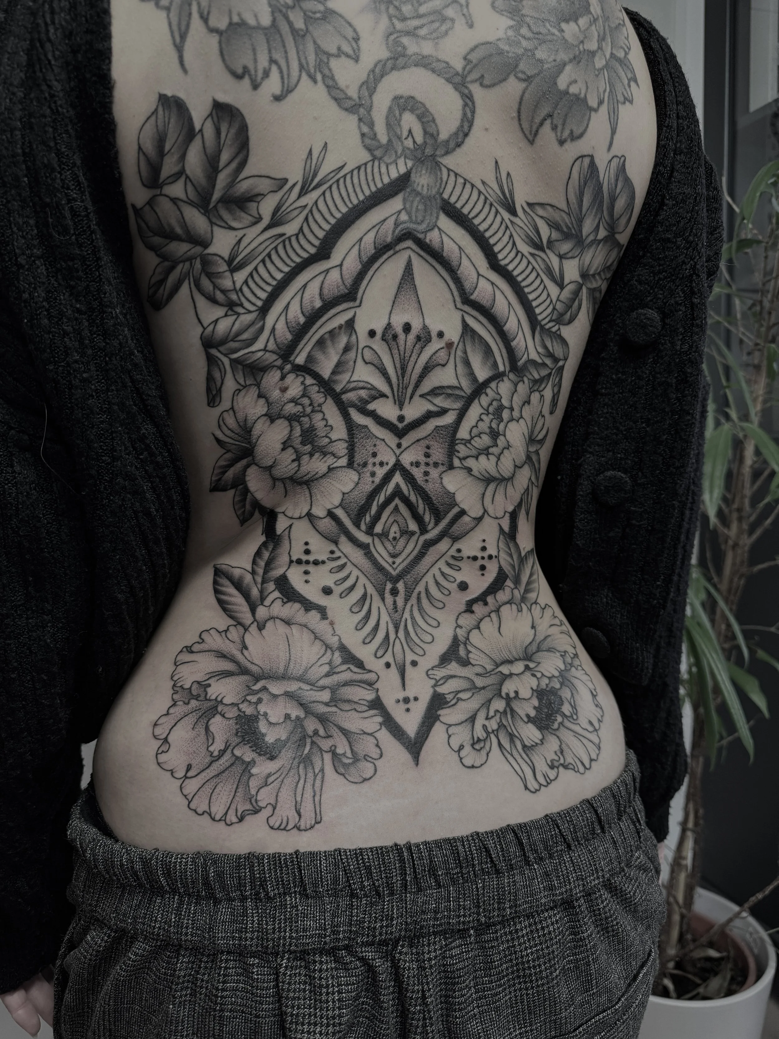 Schwarze Tätowierung mit Blumen, Blättern und geometrischen Mustern auf dem Bauch und Brust einer Person.