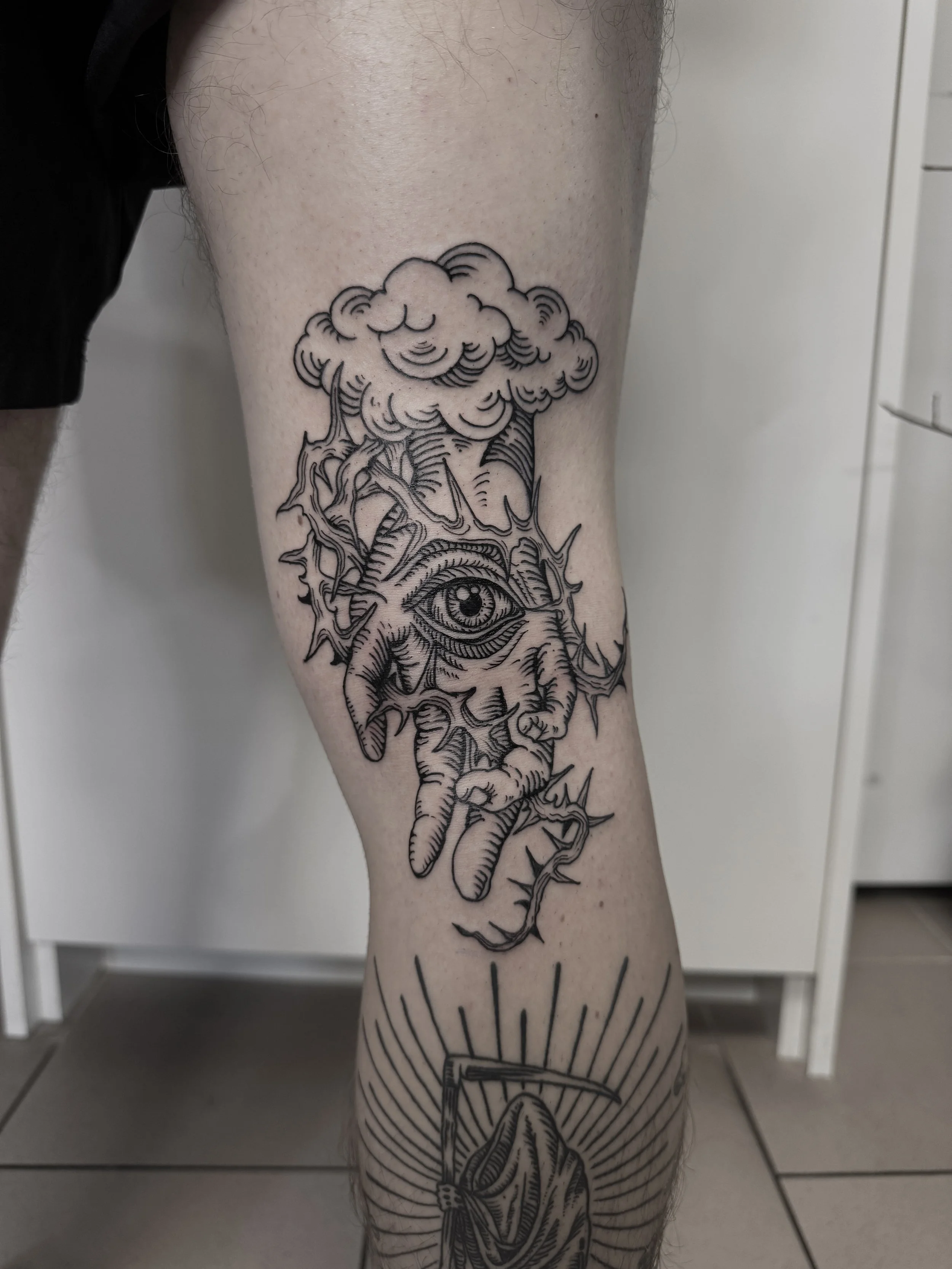 Schwarz-weiß-Tattoo auf dem Bein mit einer Hand, einem Auge in der Hand, einer Wolke und einer Sense mit Sackleinen.
