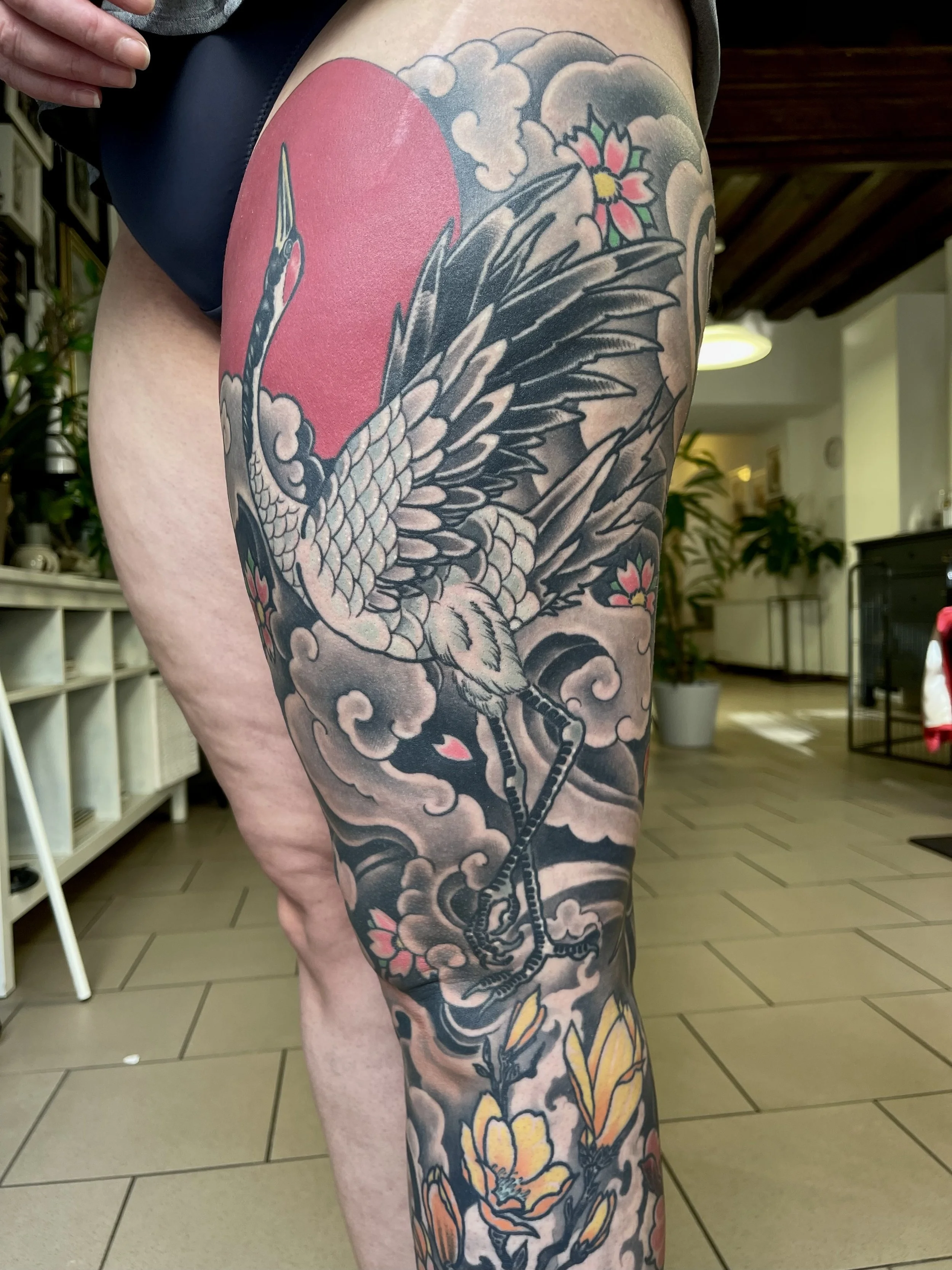 Tattoo eines Kranichs mit Blumen, Wolken und Mond auf dem Oberschenkel.