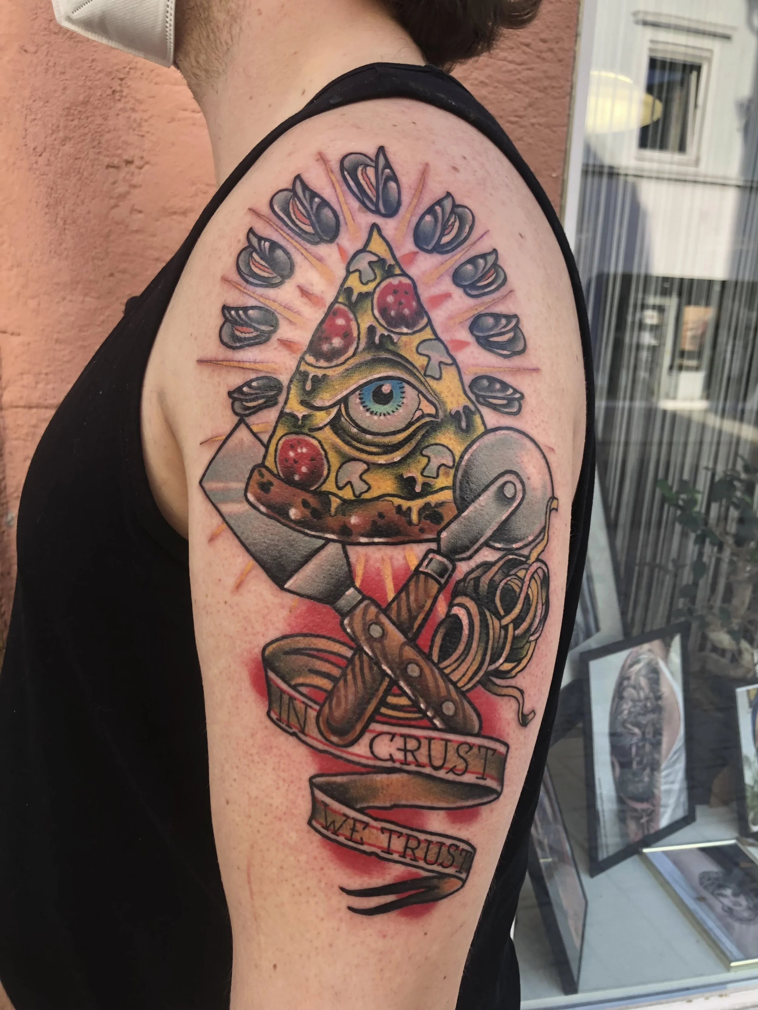 Tattoo auf dem Oberarm einer Person, zeigt eine Pizza mit einem Auge in der Mitte, umgeben von einem halbkreisförmigen Arrangement von Pilzen, unter einem Messer und einem Pizzaschneider, mit einem Banner mit den Worten 'CRUST' und 'WE TRUST'.
