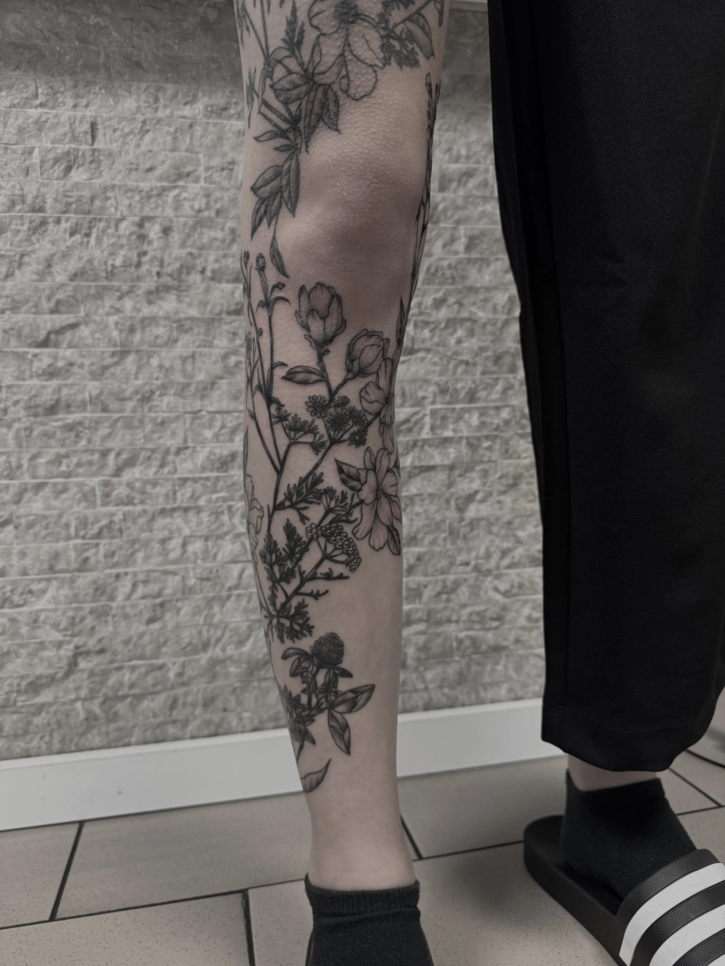 Ein Bein mit einer schwarzen Blumen-Tattoo, das auf einem Fußboden steht, nah am Wand.
