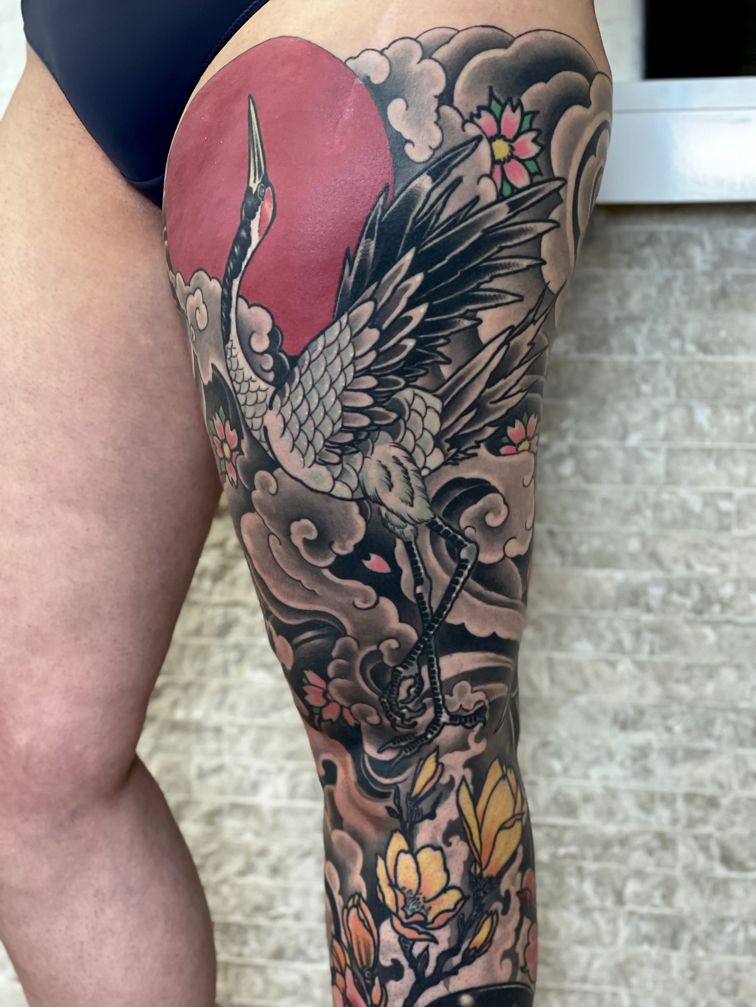Tattoo eines Kranichs mit schwarzen Flügeln, roten Sonne im Hintergrund, Wolken und Kirschblüten auf dem Oberschenkel.