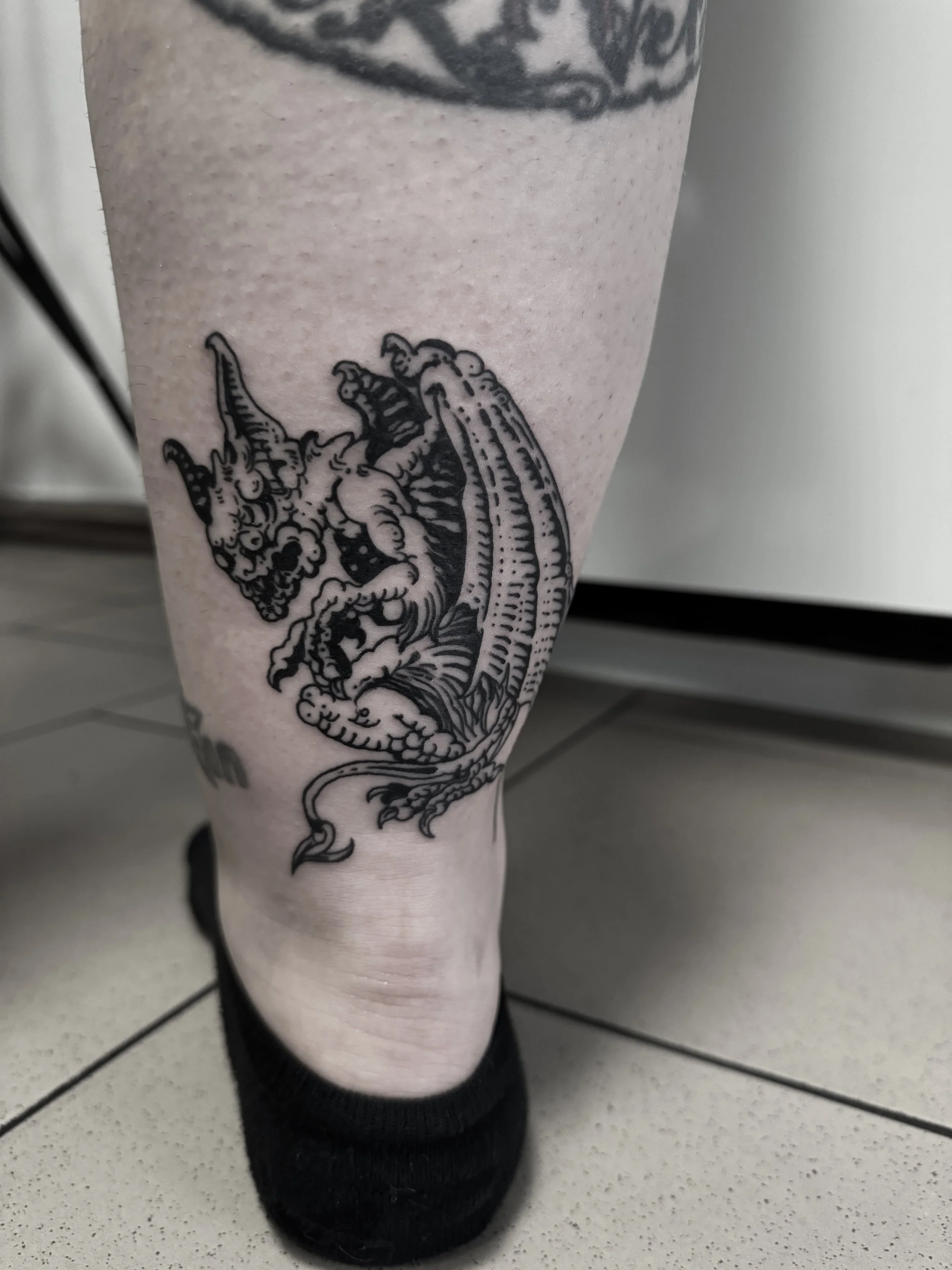 Detaillierte schwarze Tätowierung eines Drachen am Unterschenkel einer Person.