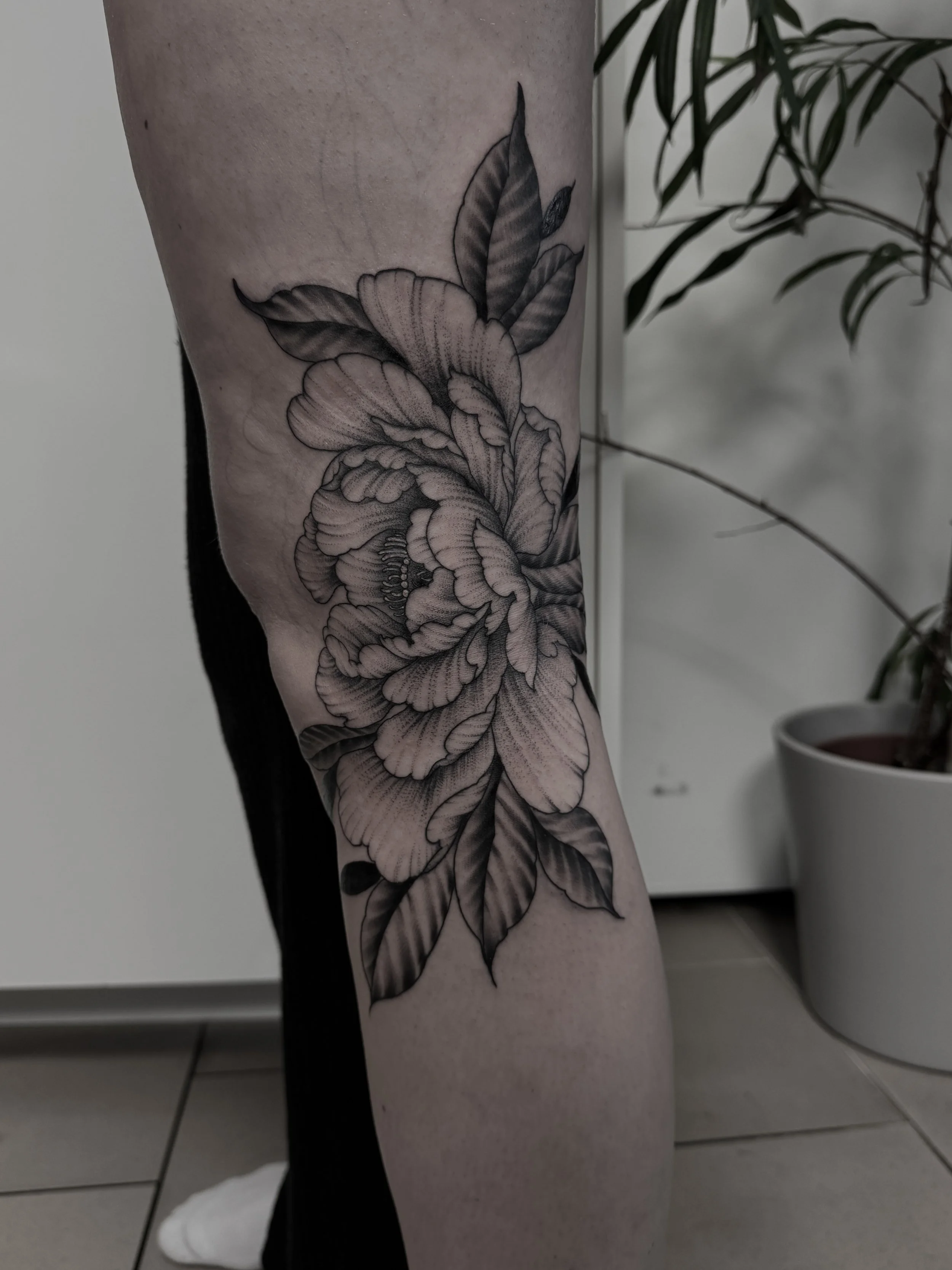 Schwarz-weiß Tattoo einer großen Blume mit Blättern am Bein.