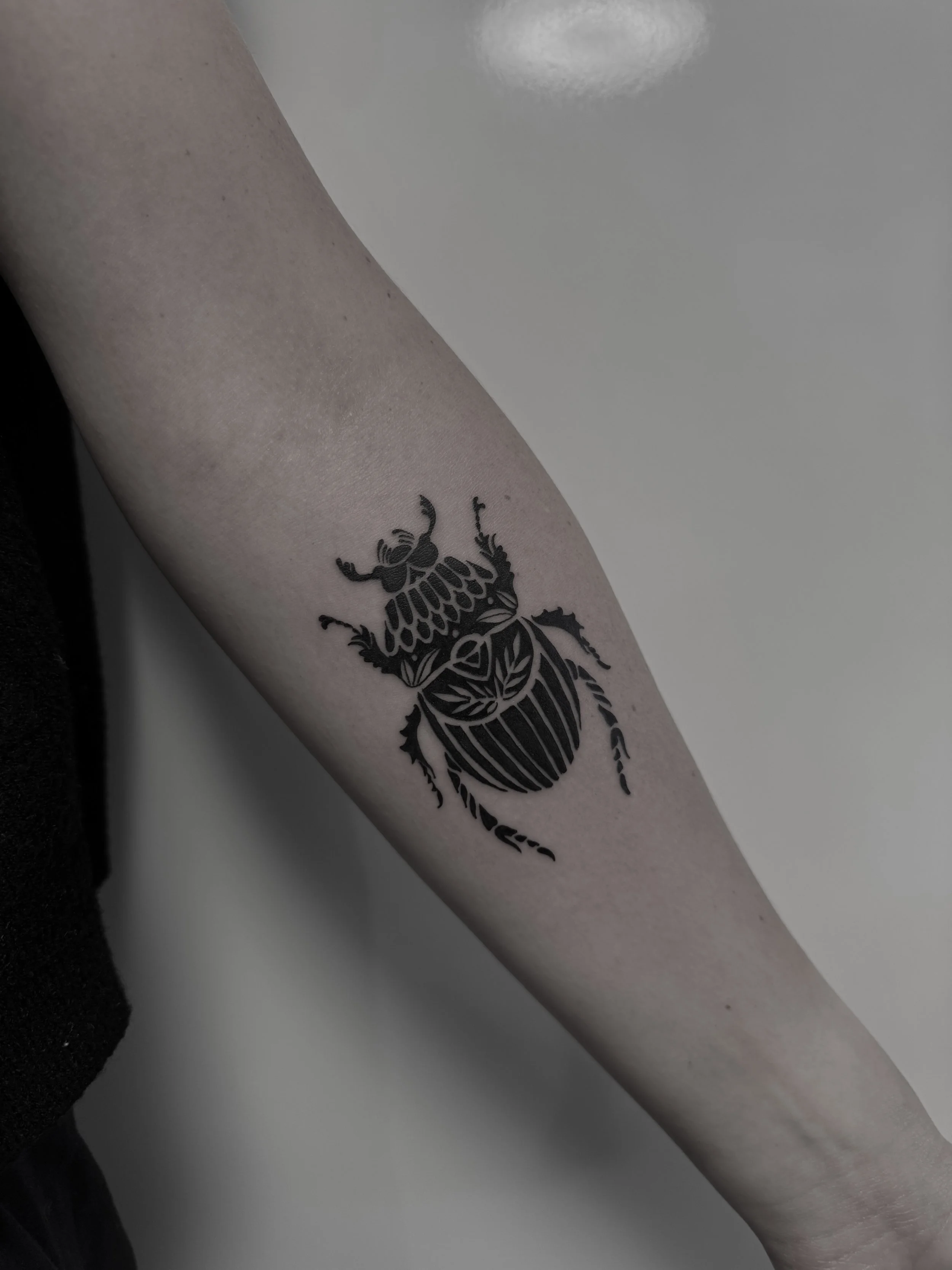 Schwarze Tätowierung eines Käfers auf dem Unterarm einer Person.