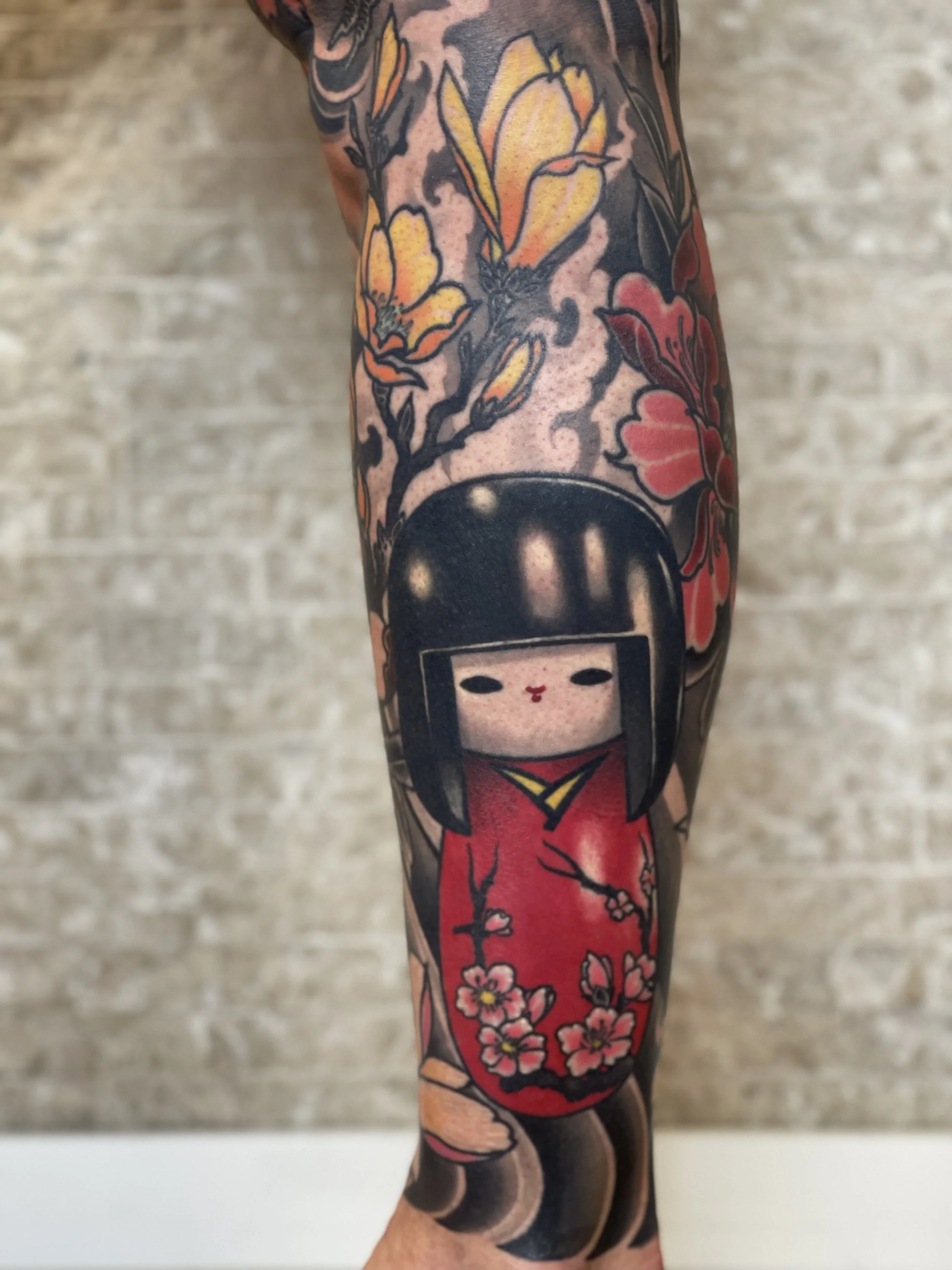 Tattoo eines Kokeshi-Mädchens, umgeben von Kirschblüten und Blumen auf dem Arm.