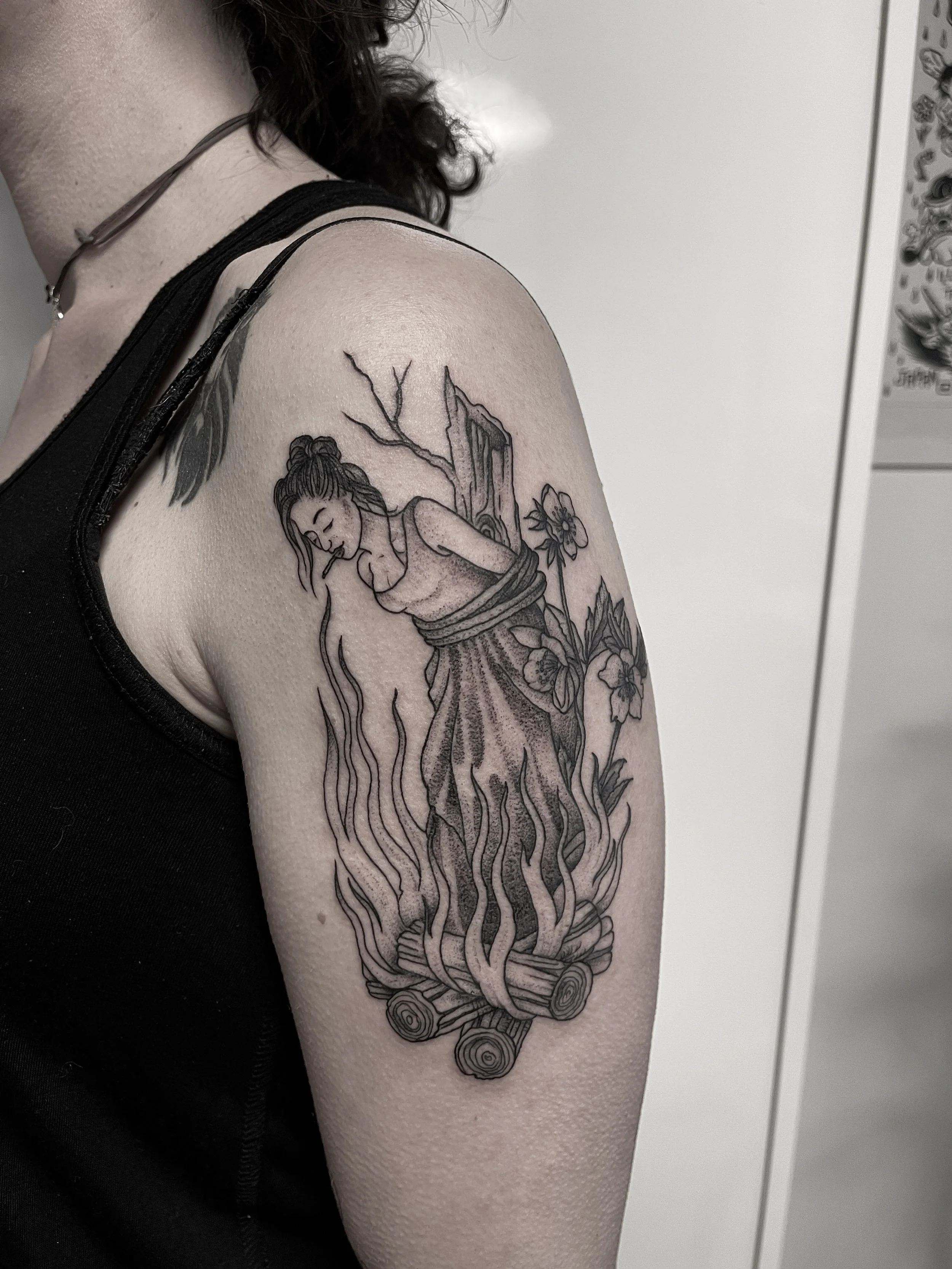 Tattoo eines Mädchens in einem abgeknoteten Kleid, das auf brennenden Holzstämmen und Blumen steht.