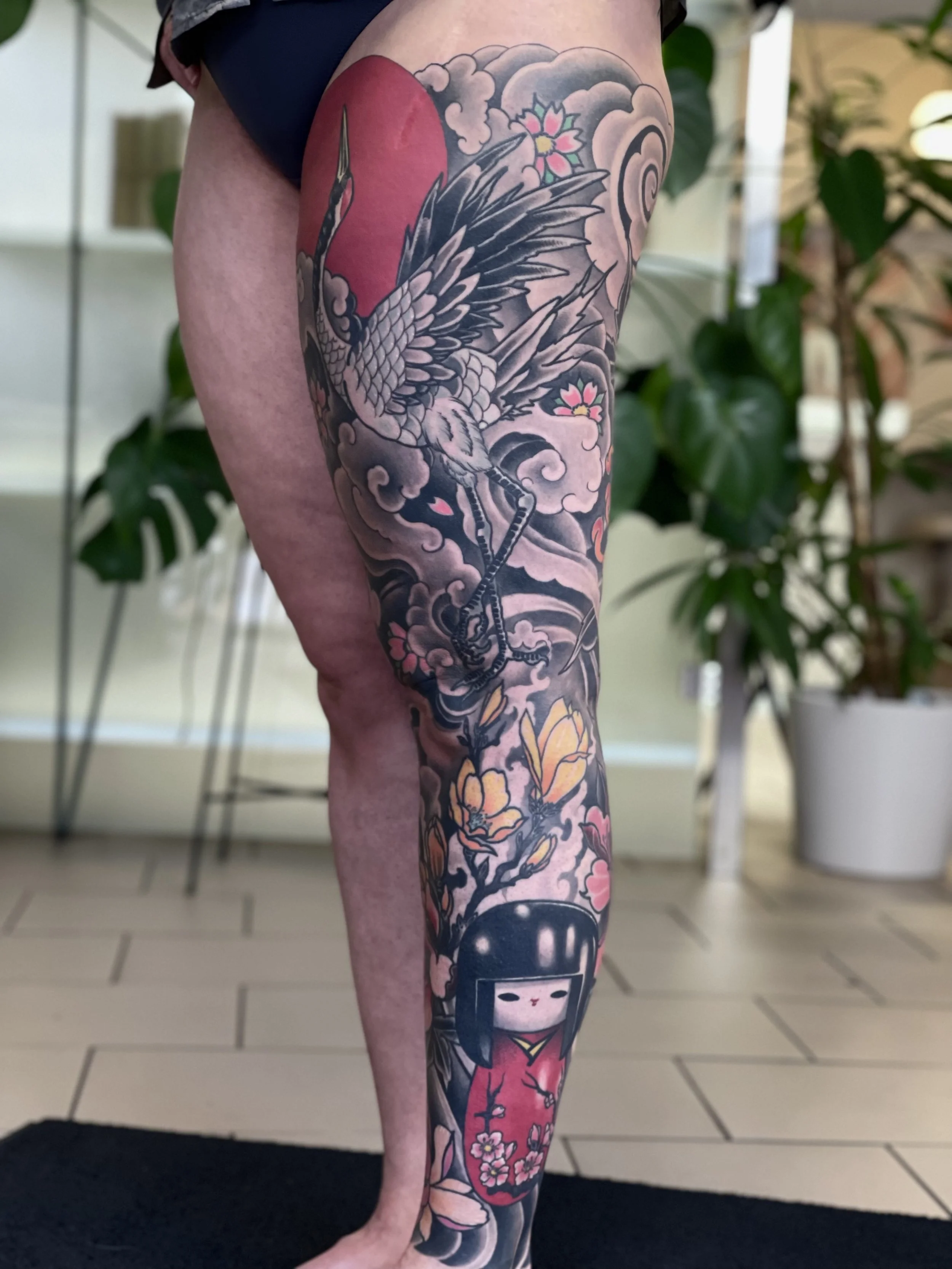 Tattoo eines Kranichs mit blühenden Kirschbäumen auf dem Bein, darunter ein Kokeshi-Püppchen.