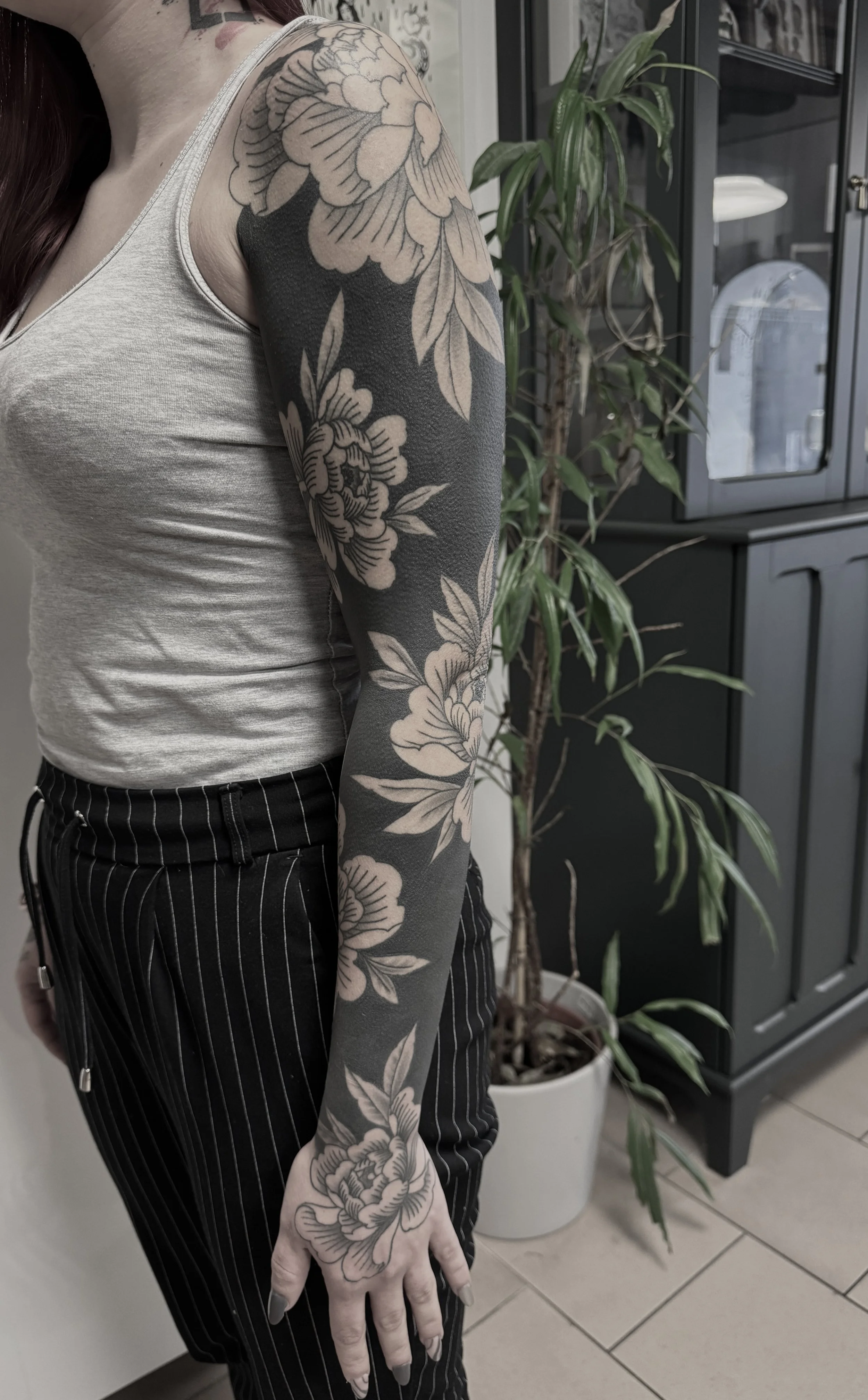 Nahaufnahme eines Arms mit schwarzen und grauen Blumen-Tattoo, die bis auf die Hand reichen, neben einer Pflanze in einem schwarzen Schrank. Person trägt ein hellgraues Tanktop und schwarze, weiß gestreifte Hose.
