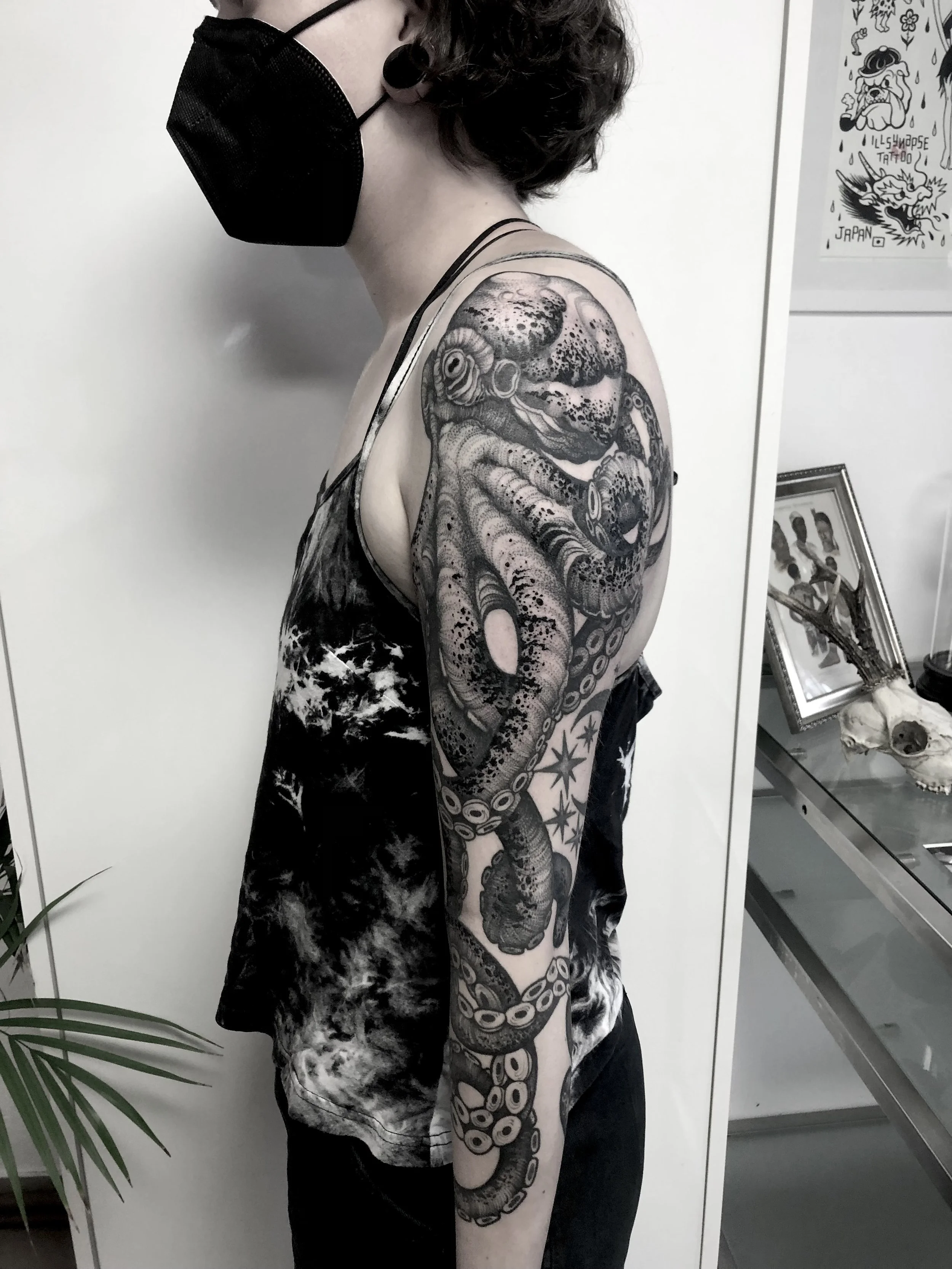 Frau mit kurzen dunklen Haaren, schwarzer Gesichtsmaske und umfangreichem schwarzen Tätowierung auf dem linken Arm und Schulter, trägt ein schwarzes Tanktop und schwarze Hosen, steht seitlich vor einer weißen Wand mit Kunstwerken