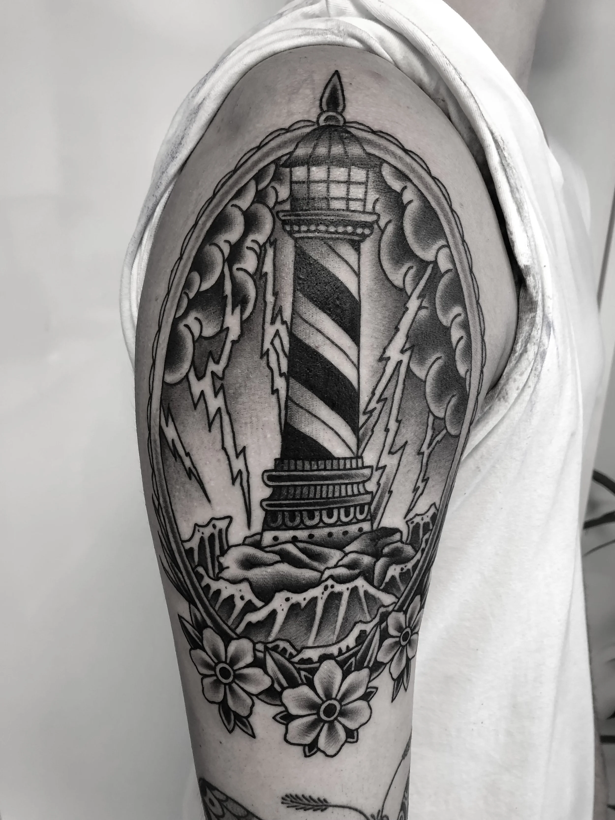 Schwarz-weiß-Tätowierung eines Leuchtturms mit Blitzschlägen und Blumen auf dem Oberarm.