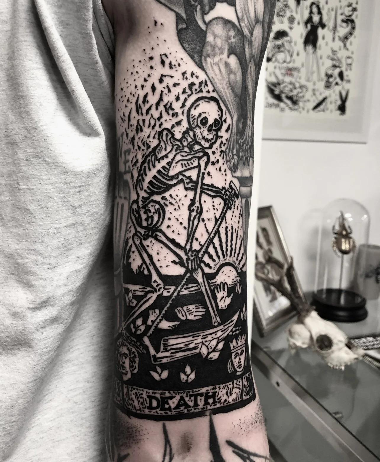 Schwarze Tätowierung eines Skeletts auf dem Oberarm, das auf einer Waage steht und den Sonnenaufgang im Hintergrund zeigt, mit dem Wort "Death" am unteren Rand.