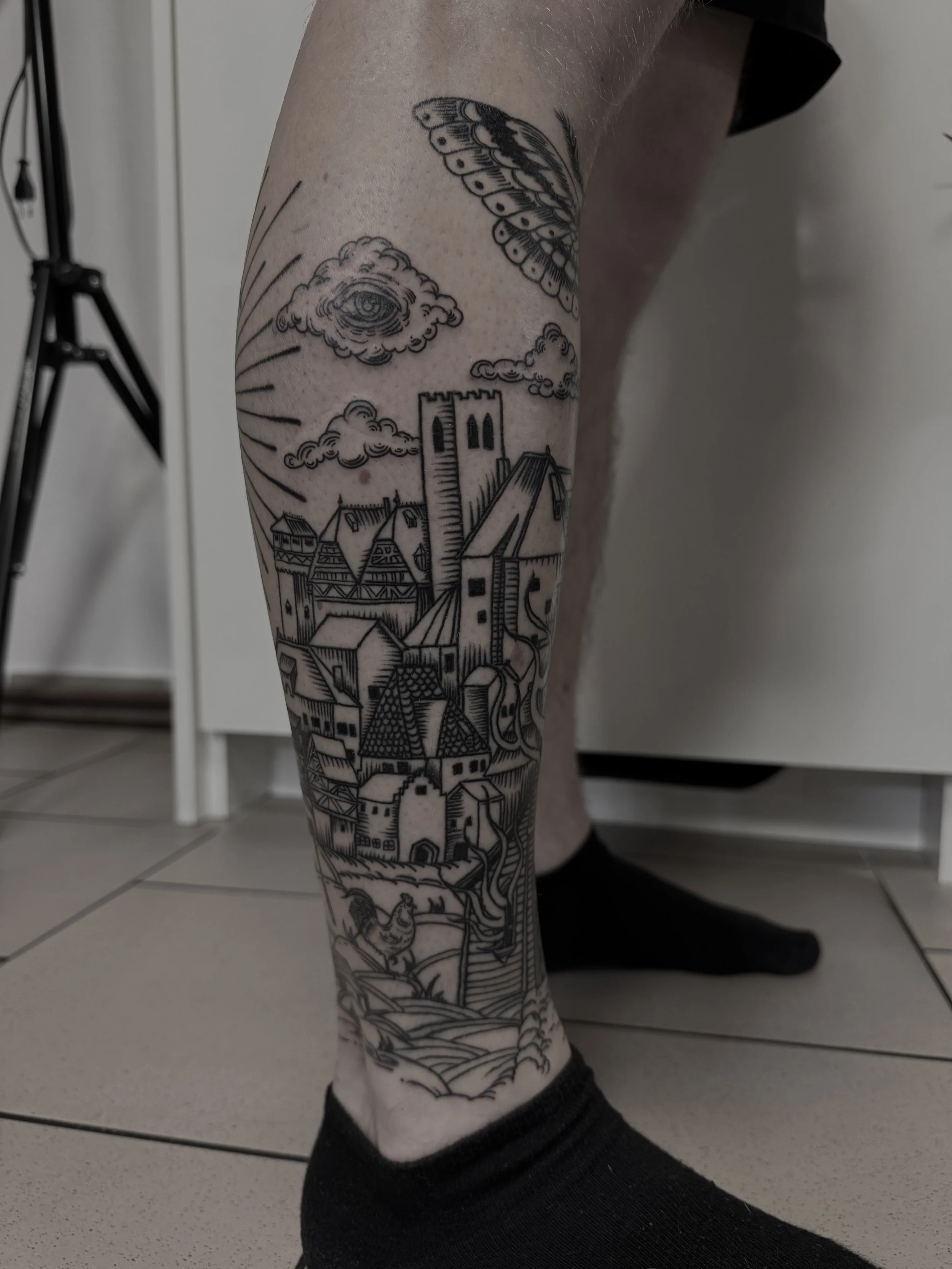 Schwarz-weiß-Tattoo eines Dorfes mit Türmen, Häusern und Hügel auf dem Unterschenkel, mit Wolken und einem großen Auge in der Wolke und einem Schmetterling am oberen Rand.