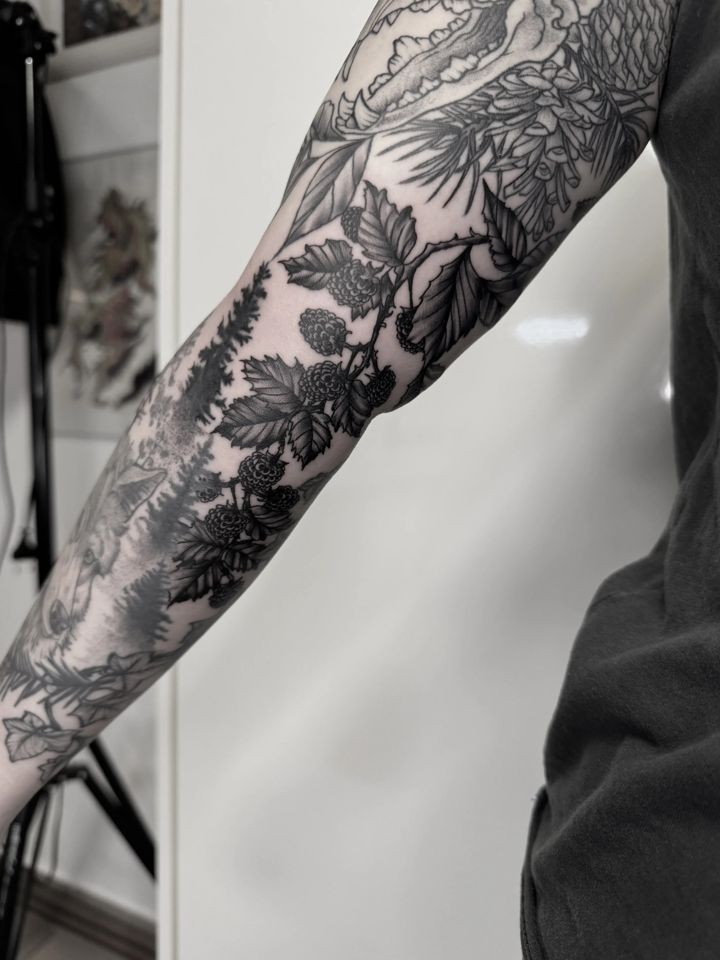 Schwarze Tätowierungen mit Pflanzen- und Naturmotiven, darunter Beeren und Blätter, auf dem ausgestreckten Arm einer Person.