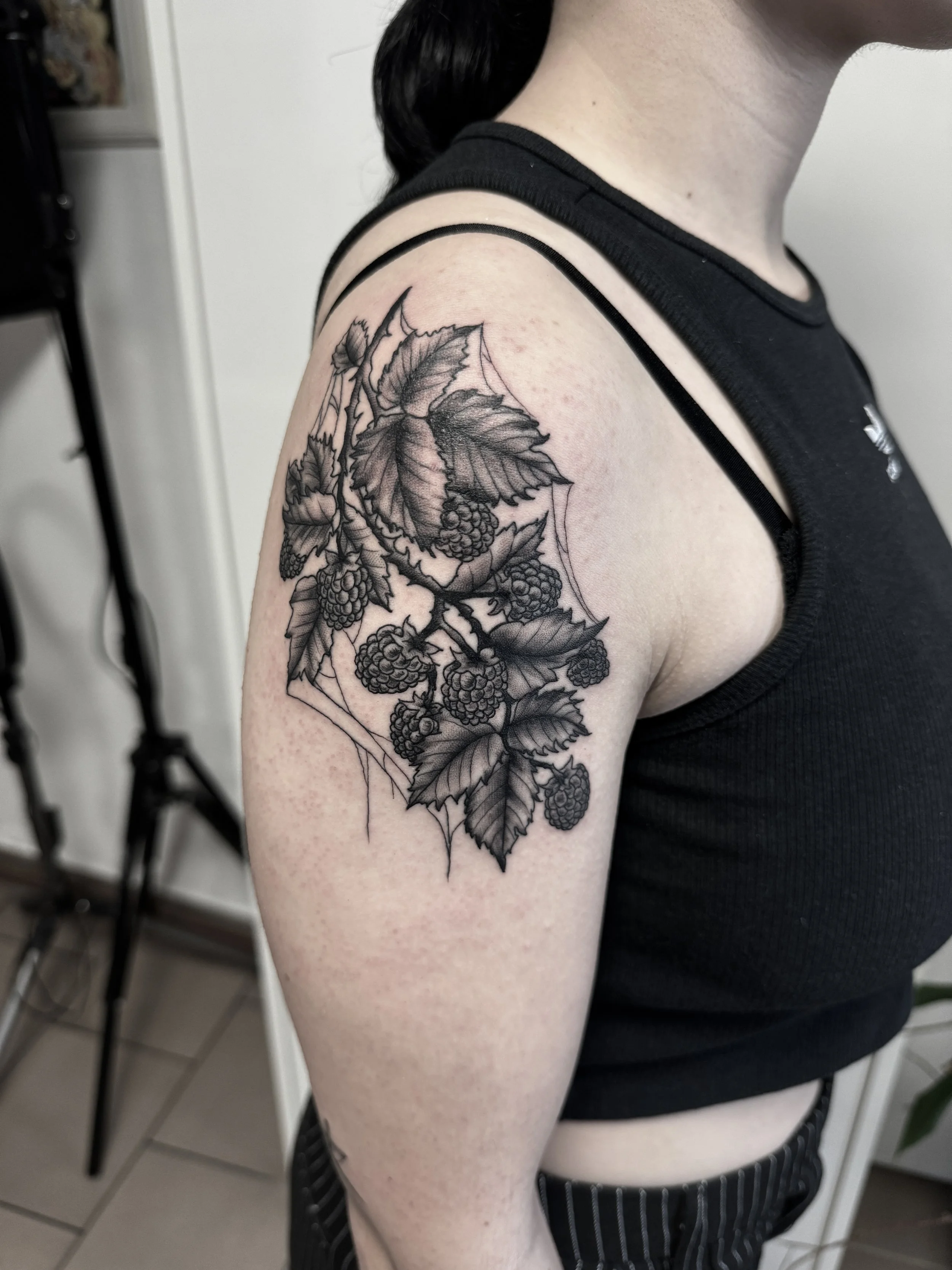 Schwarz-weiß-Tattoo einer Brombeer-Pflanze auf dem Oberarm einer Frau mit schwarzen Haaren, trägt einen schwarzen ärmellosen Oberteil.