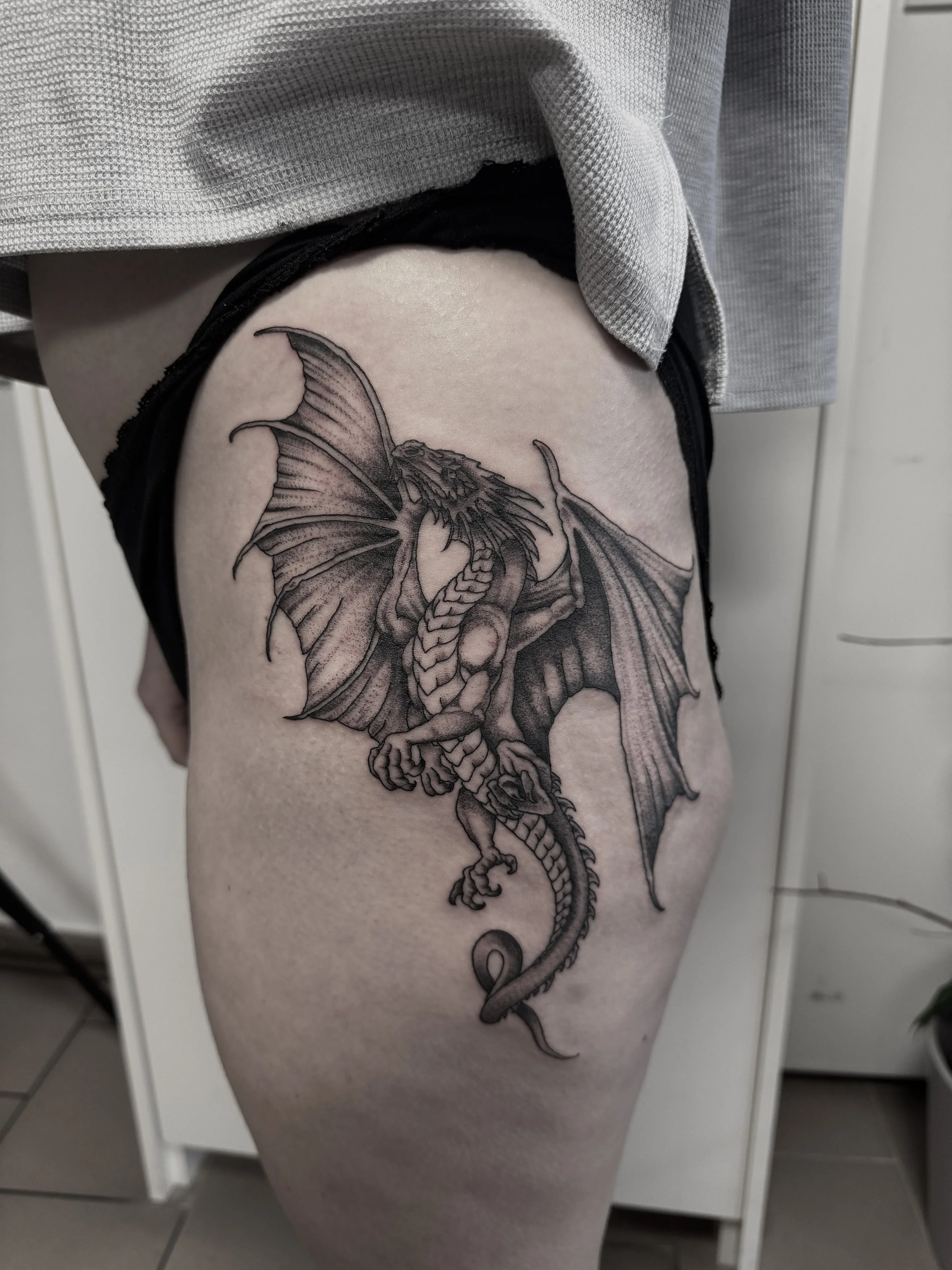 Schwarze Tätowierung eines Drachen auf Oberschenkel. Der Drache hat Flügel, gehärtete Gliedmaßen und einen langen, geschwungenen Schwanz.