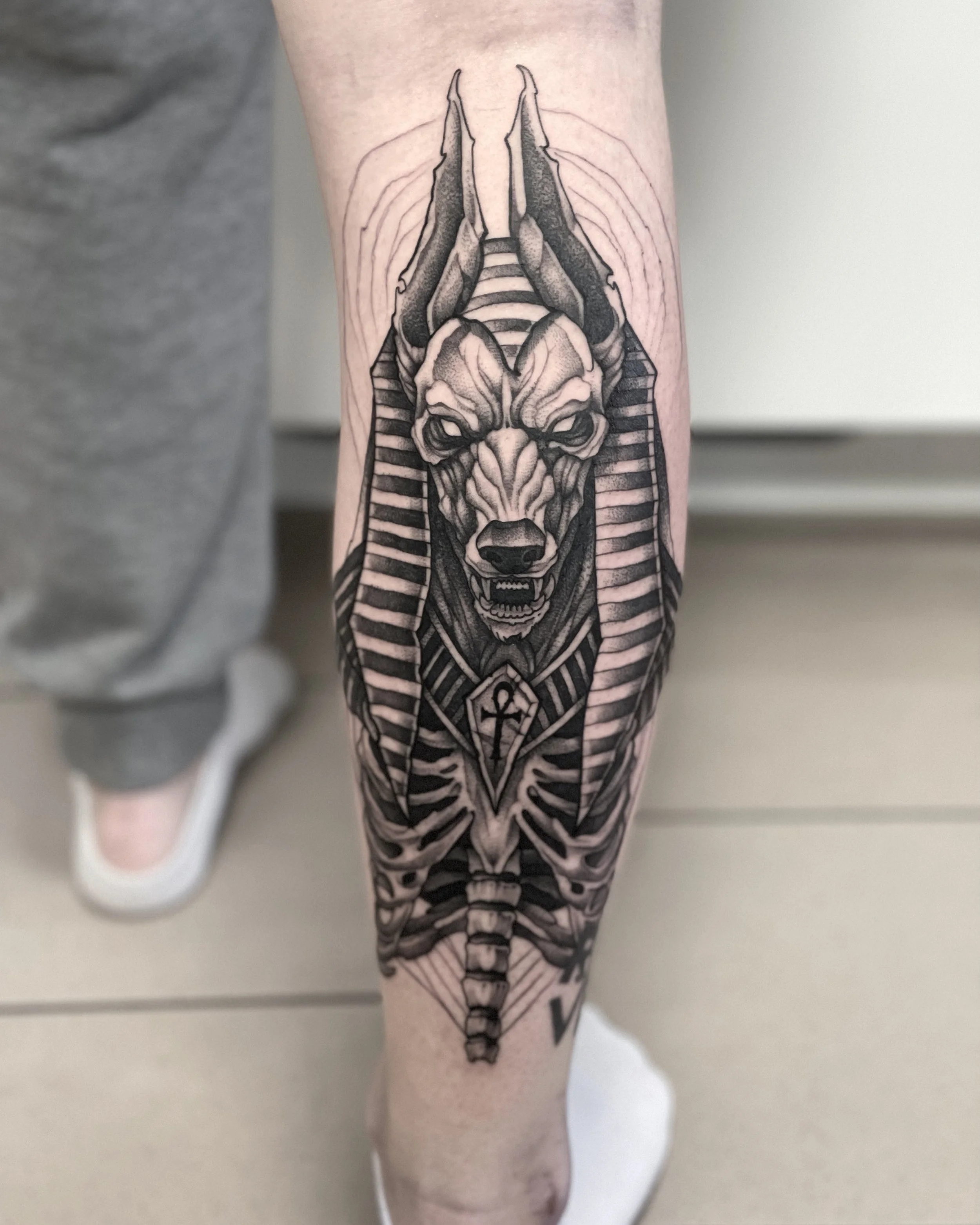 Schwarze Tätowierung eines Hundes mit Ägyptischer Pharaonen-Kleidung auf dem Unterarm.