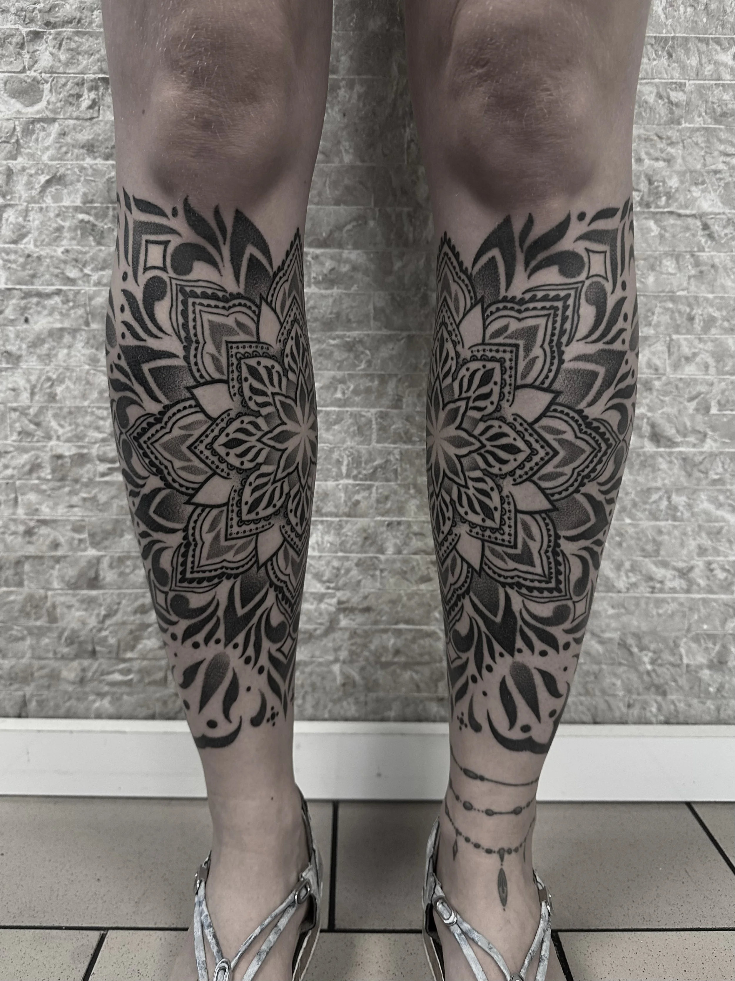 Beine mit großen schwarzen Mandala-Tattoos, die von den Knien bis zu den Füßen reichen, vor einer Steinmauer in High Heels.
