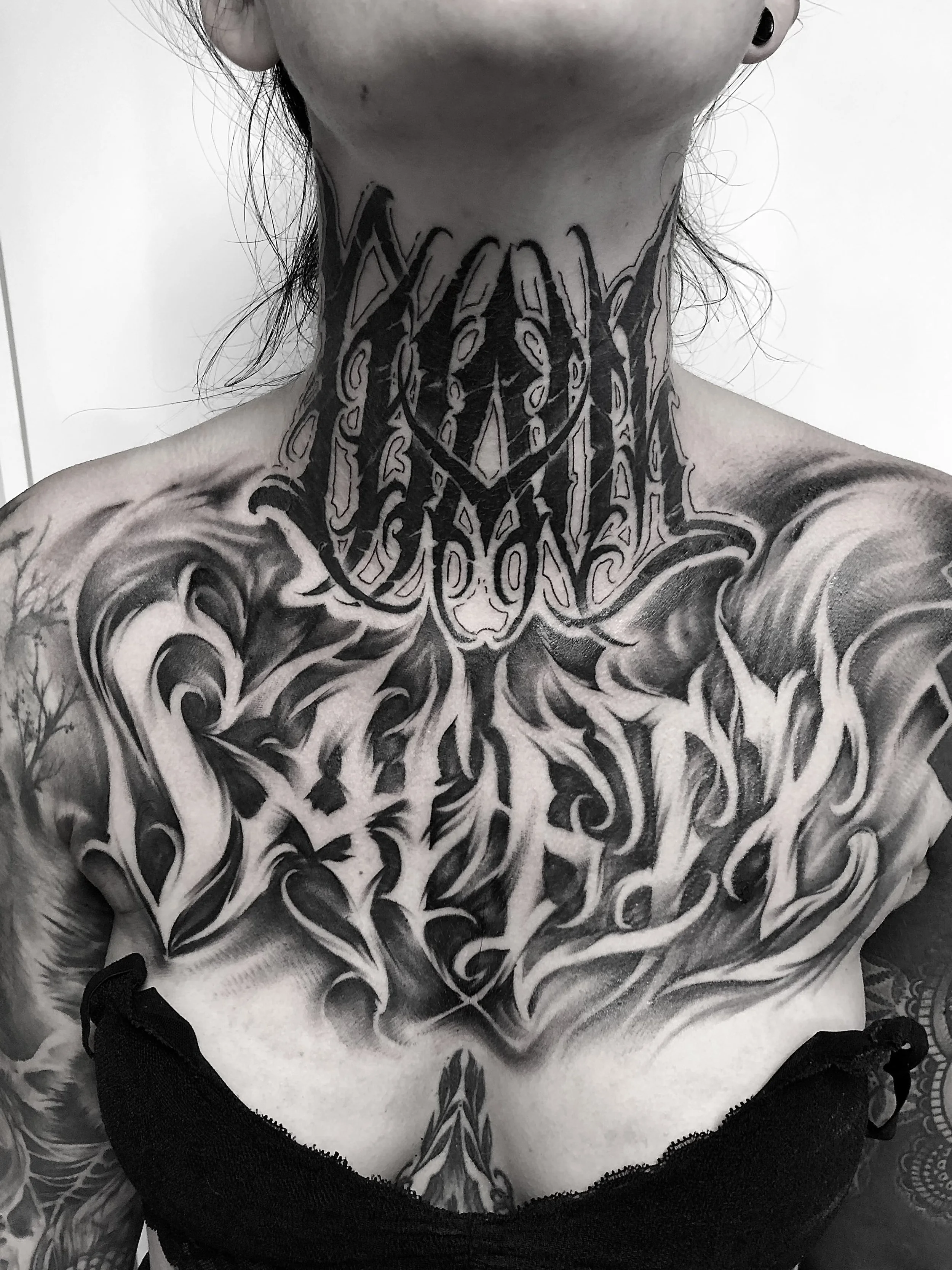 Schwarze Tätowierungen auf Hals, Brust und Schultern einer Person, mit komplexen, schwarzen Linien und Mustern in dunklem Stil.
