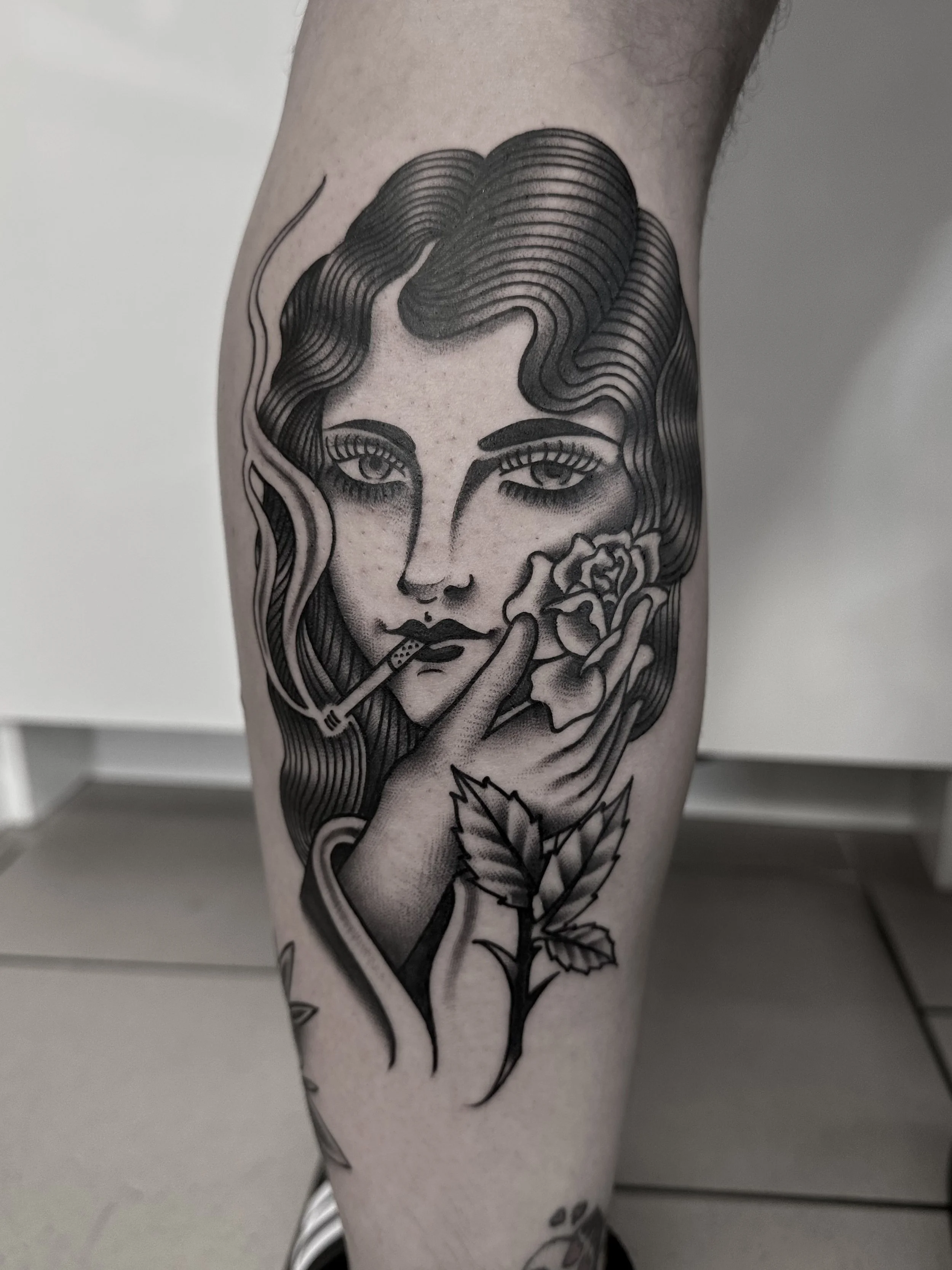 Schwarz-Weiß-Tattoo einer Frau mit welligem Haar, die eine Zigarette hält, eine Rose an ihrem Gesicht und Blätter. Der Stil ist line-art mit Schattierungen.