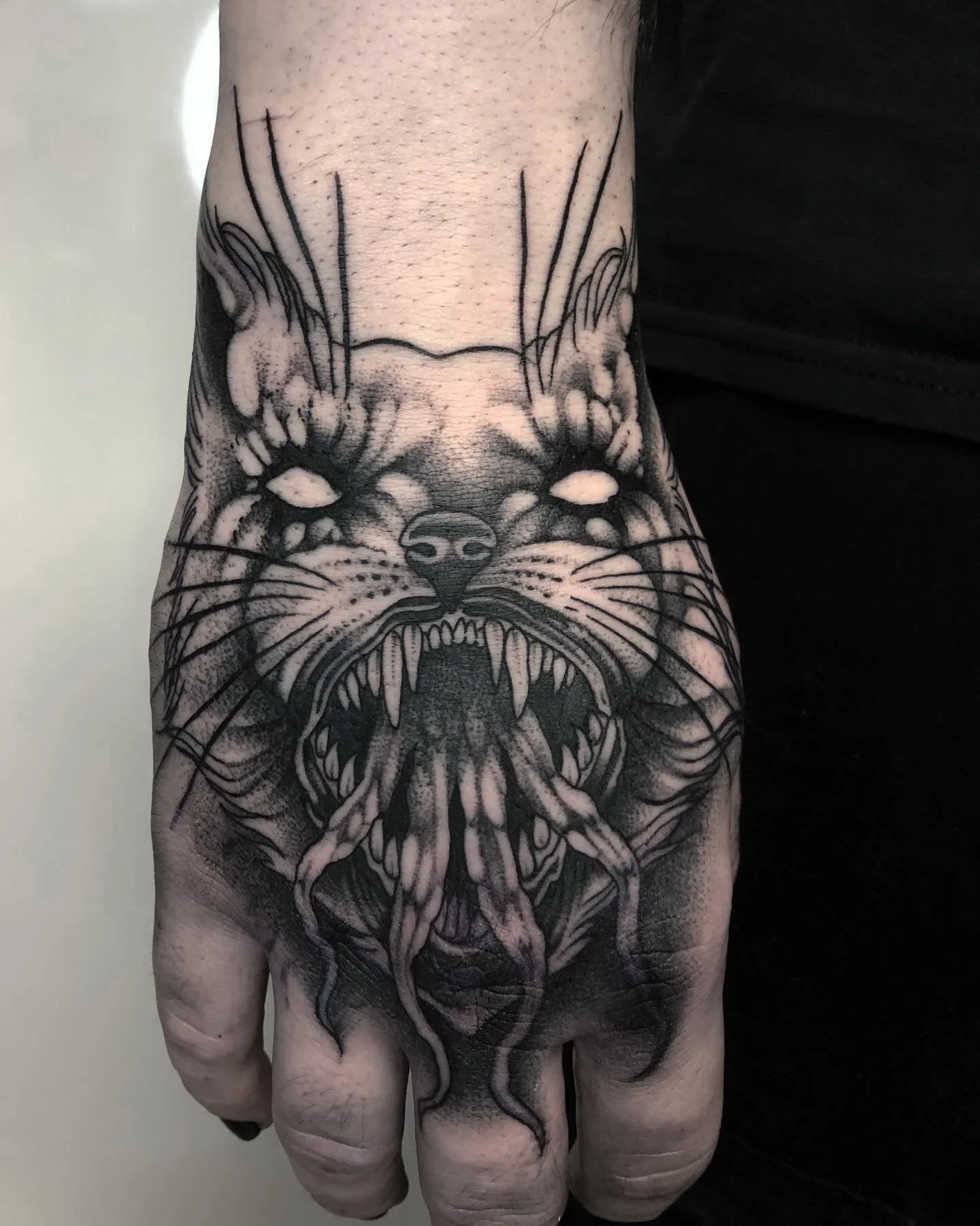 Schwarze Tätowierung eines wütenden Katzenkopfes auf der Hand mit offenen Mäulern, Zähnen und Zunge, in realistischem Stil.