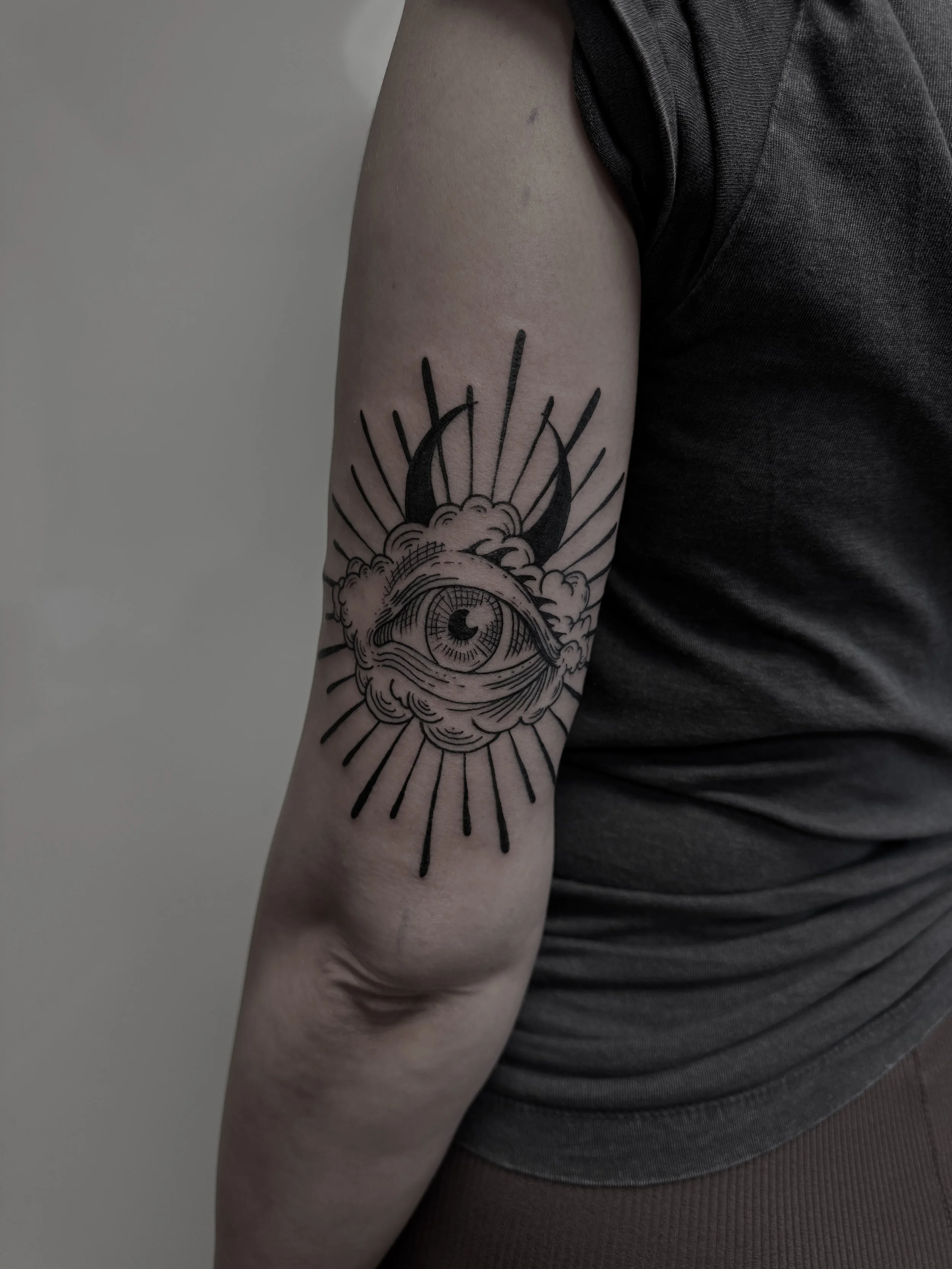 Tattoo eines Auges in einer Wolke mit Sonnenstrahlen auf dem Oberarm einer Person.