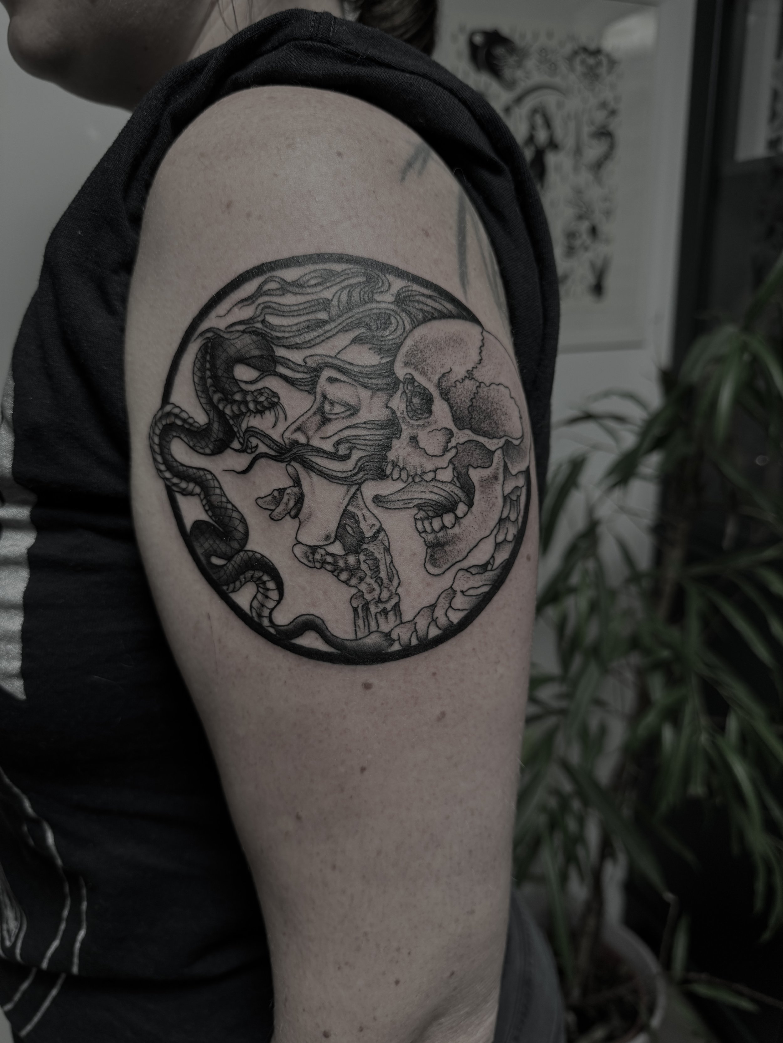 Tattoo auf dem Oberarm, das eine halb geschlossene menschliche Figur mit langen Haaren zeigt, umgeben von zwei sich gegenüberstehenden Skeletten innerhalb eines Kreises.