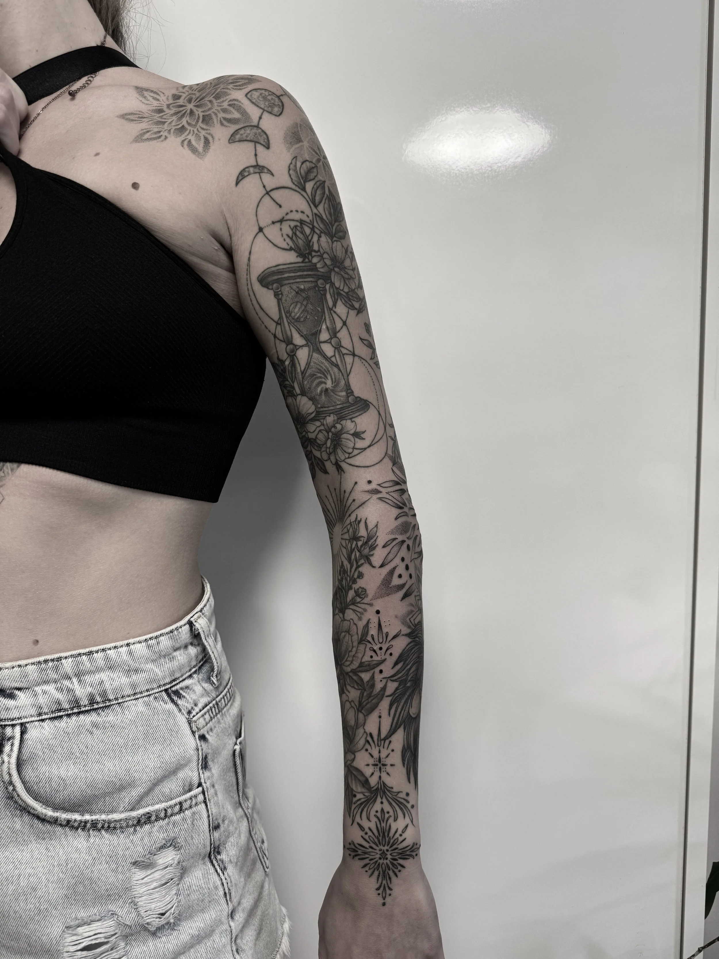 Nahaufnahme eines weiblichen Arms mit schwarzen Tattoos, darunter ein schwarzes Oberteil und hellblaue, zerrissene Jeans.