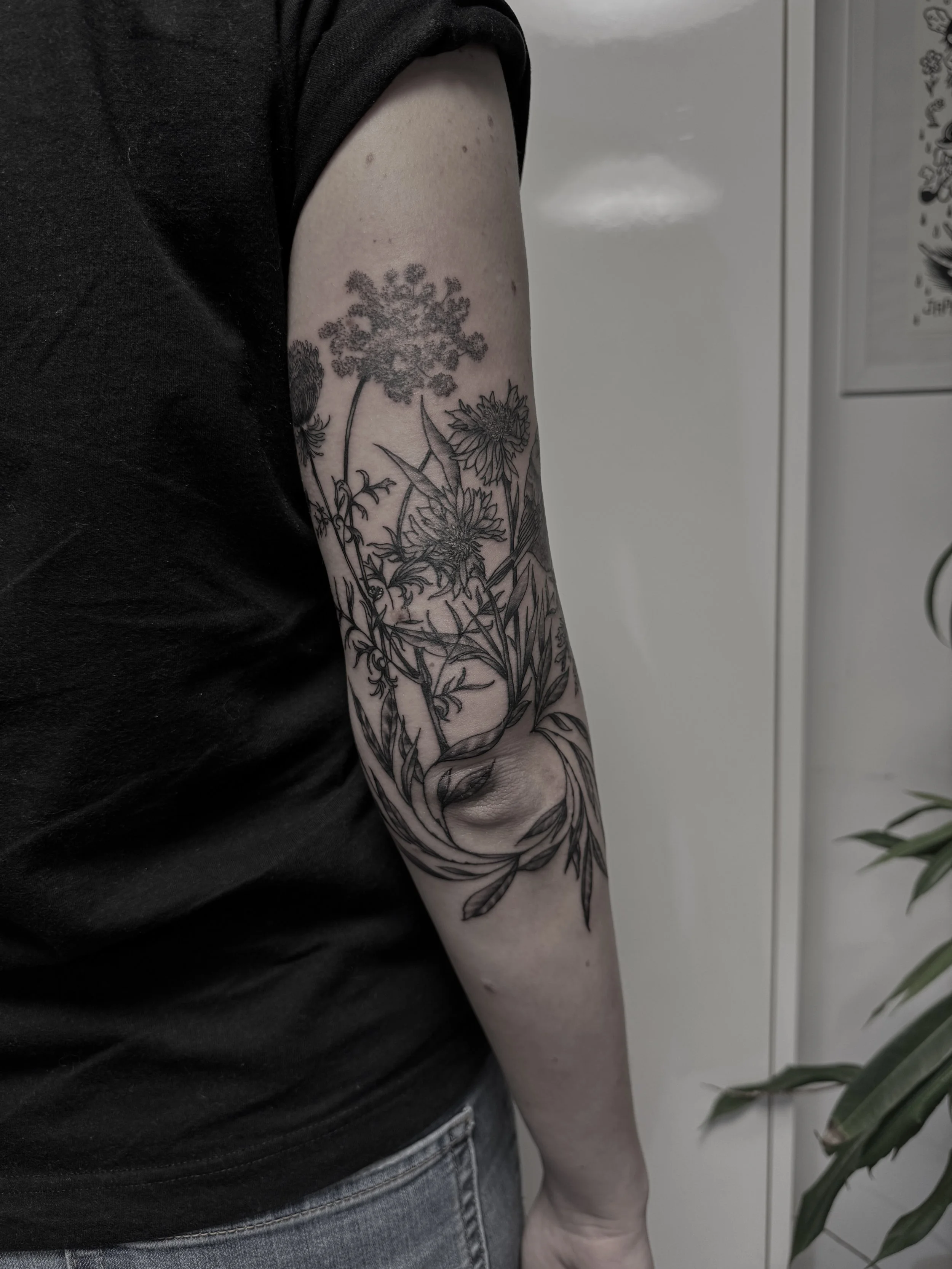 Schwarze Tätowierung mit Blumen auf dem Unterarm einer Person, die schwarze Kleidung trägt.
