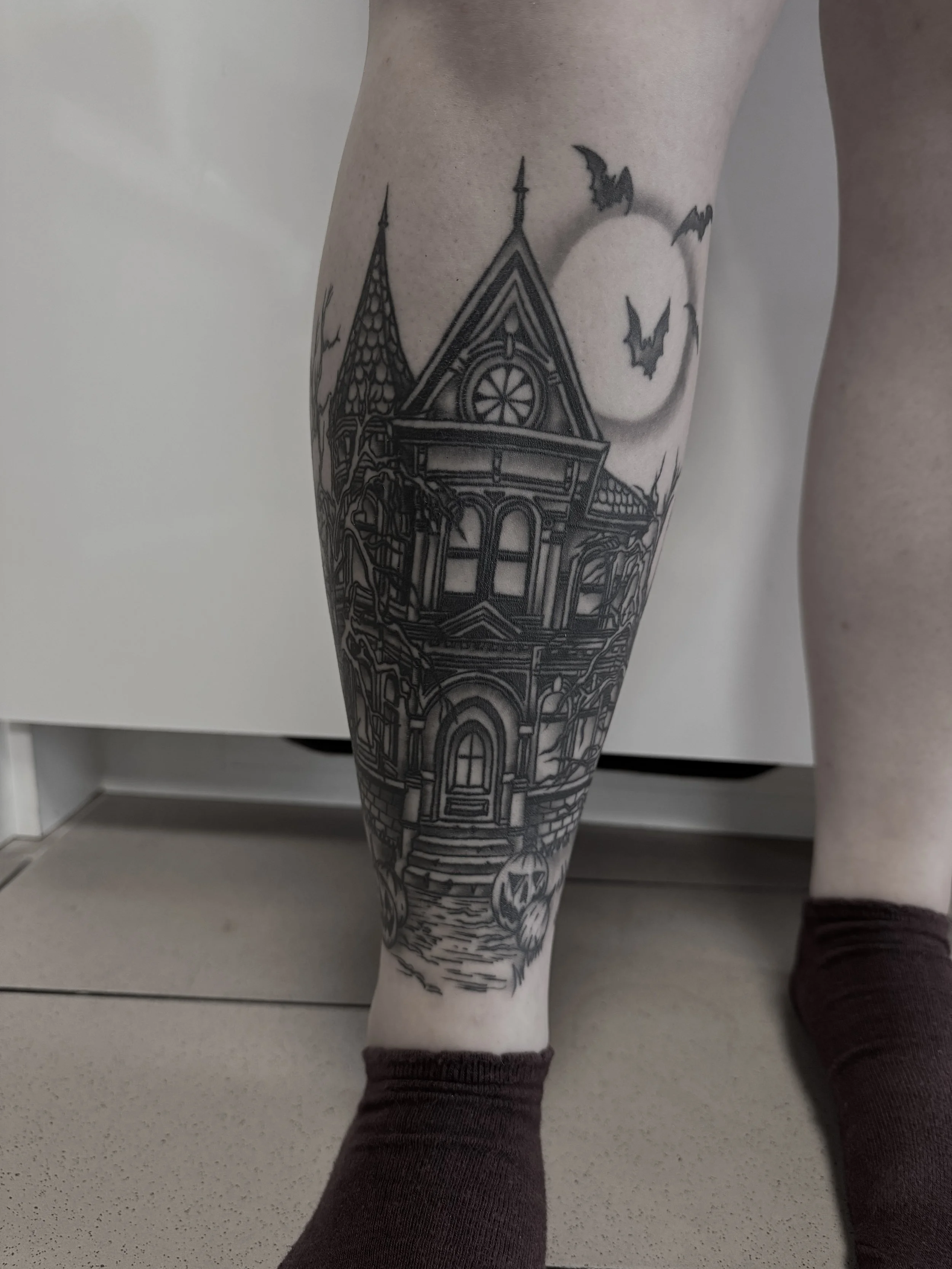 Schwarze Tätowierung eines Halloween-Hauses mit Vitamin-Vergolden auf einem Bein, vor einem Vollmond mit fliegenden Fledermäusen in der Nacht.