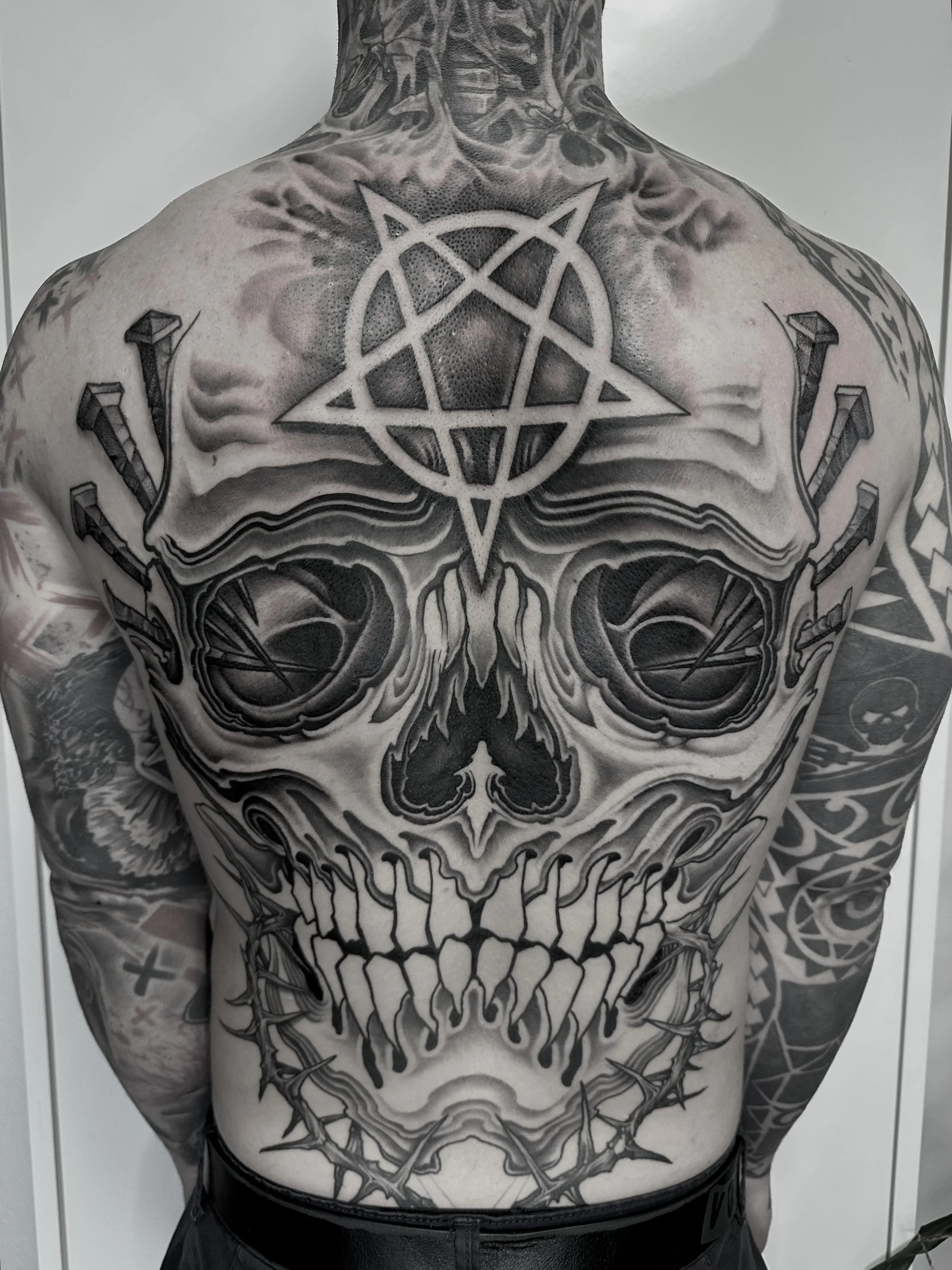 Schwarze und graue Tätowierung eines Totenschädels mit keltischem Pentagramm auf der Brust, umgeben von weiteren Designs am Arm.