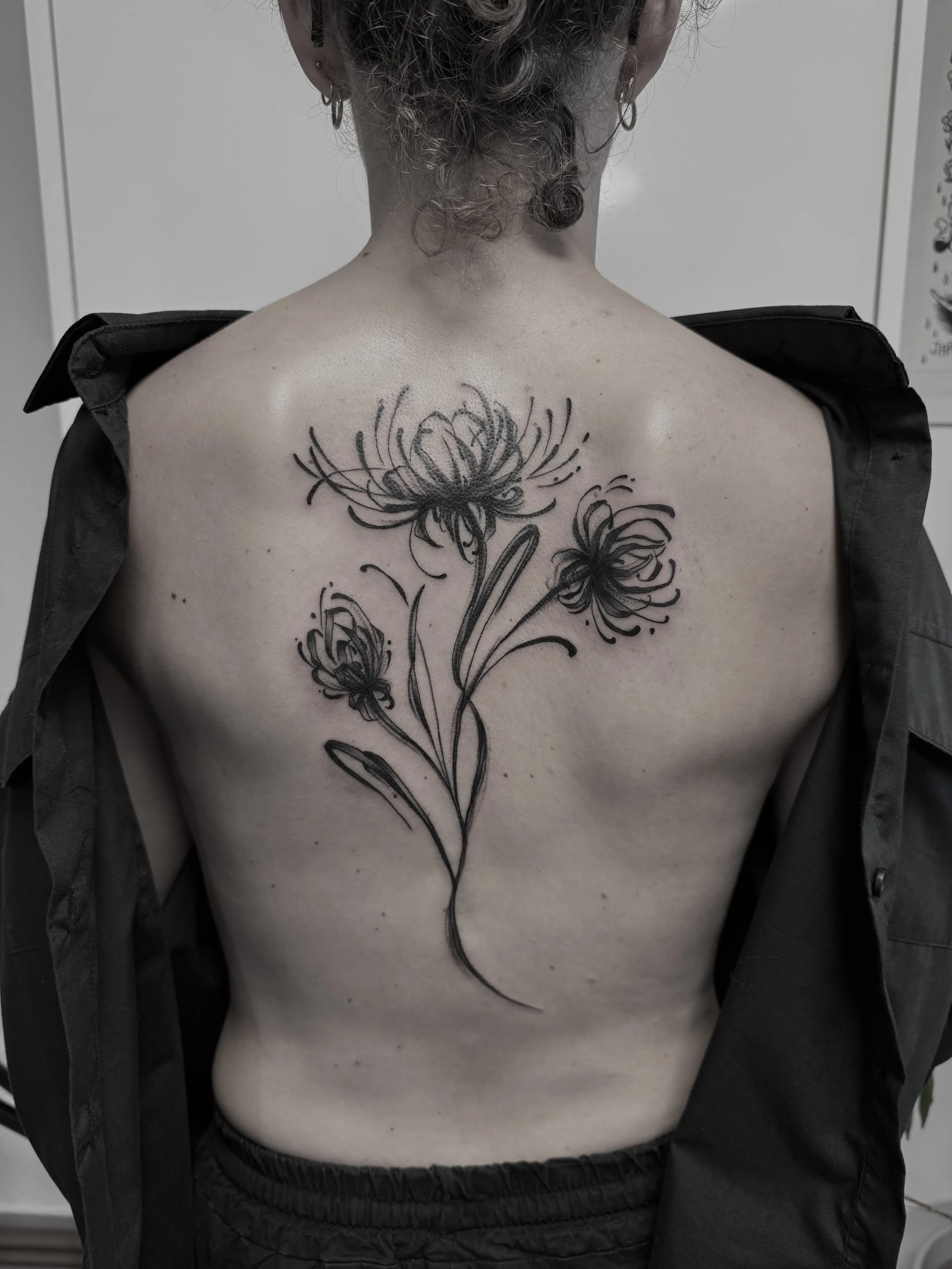 Tattoo von drei Blumen in Schwarz auf dem Rücken einer Person, die Rückenansicht, Person trägt eine schwarze Jacke.