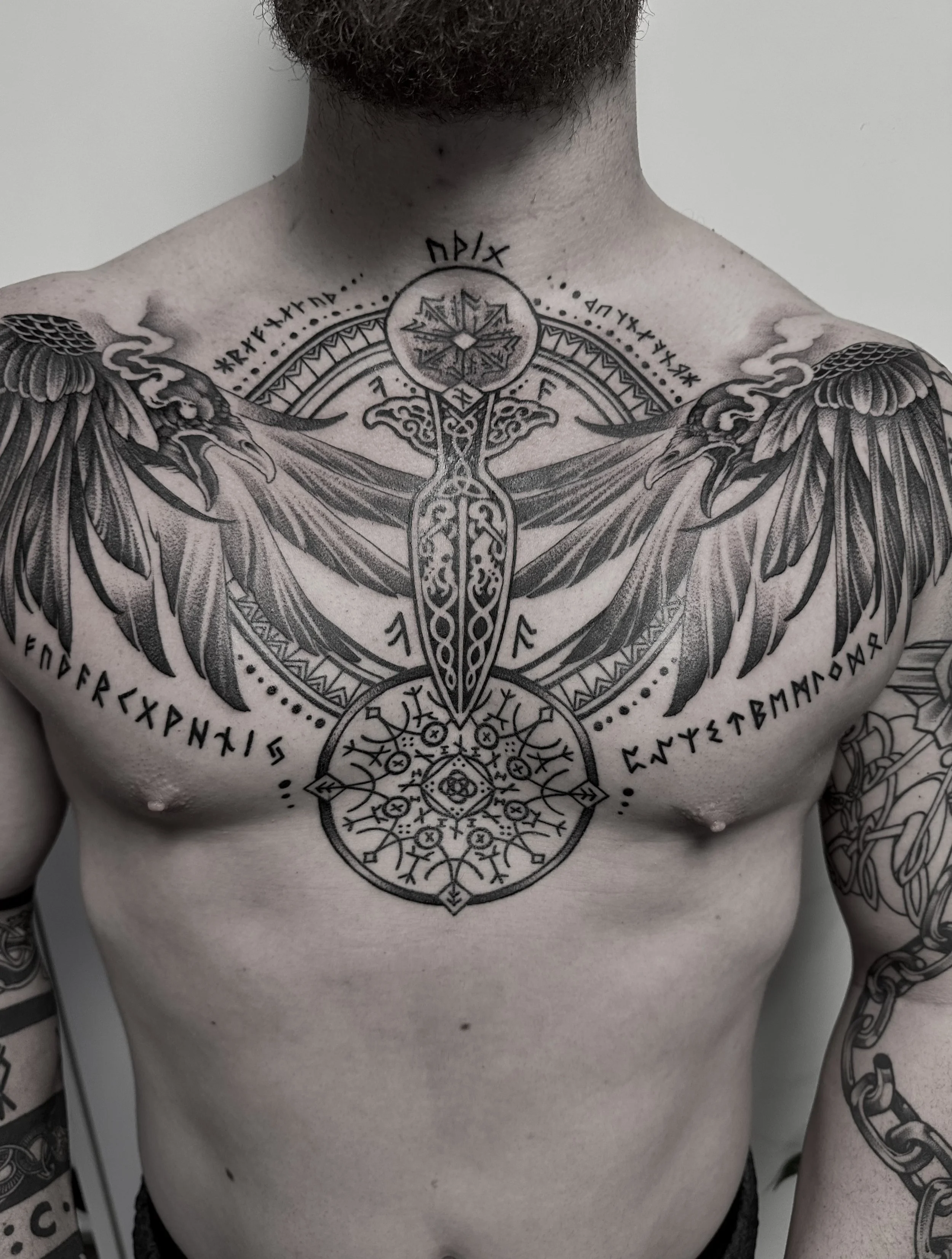 Schulter- und Brust-Tattoo eines Mannes mit nordischer Symbolik, darunter ein Kreuz, Runen und ein geometrisches Muster.