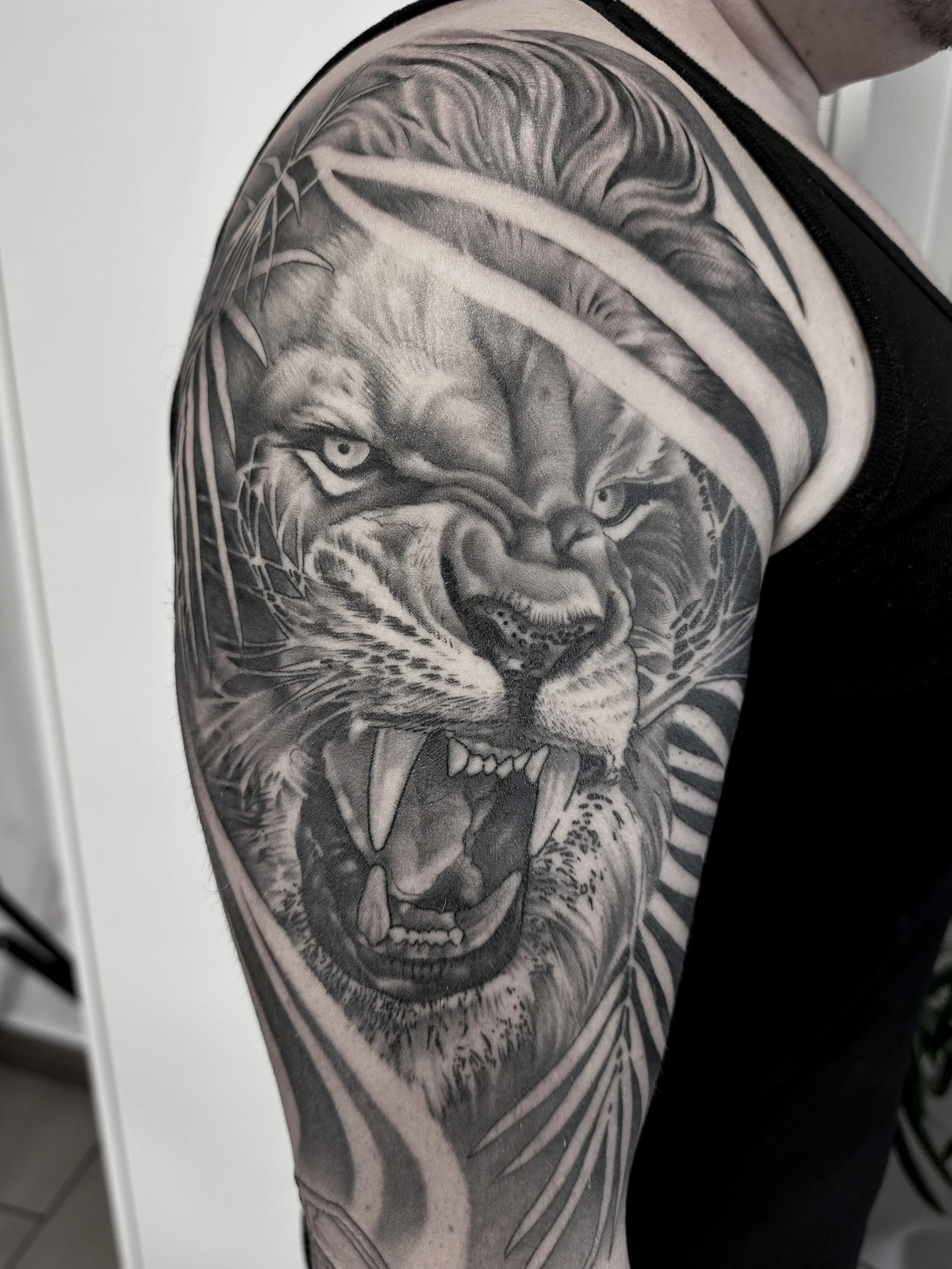 Tattoo eines löwenkopfes auf dem Oberarm, in Schwarz-Weiß, mit geöffnetem Maul und Zähnen, umgeben von Blättern und Linien.