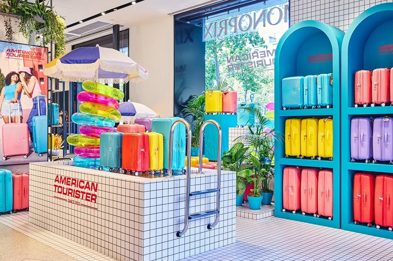 Rayons de valises colorées et objets de plage dans un magasin de tourisme, avec plantes vertes et publicité en arrière-plan.