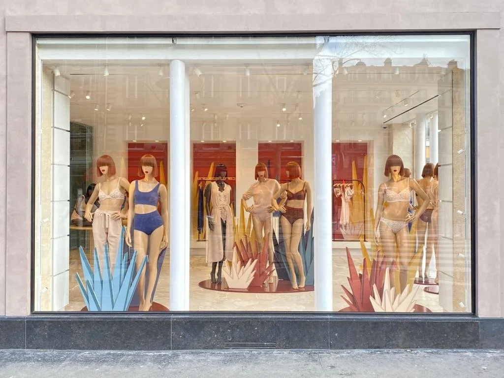 Vitrine d'un magasin de lingerie avec des mannequins portant des sous-vêtements en dentelle, entourés de décorations colorées en forme de flammes ou de feuilles dans la fenêtre.