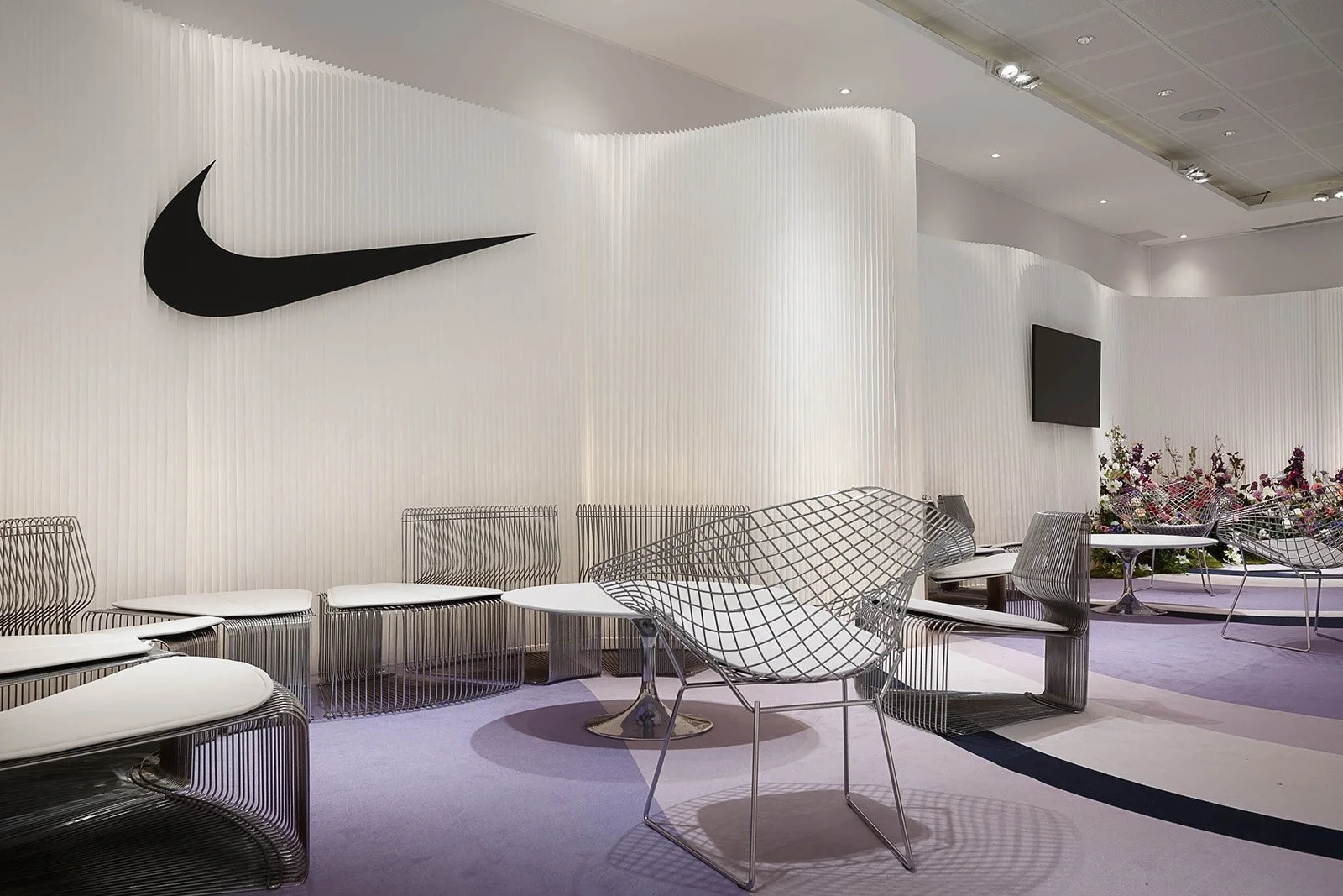 Une salle d'attente moderne avec des chaises en fil métallique et un mur blanc avec le logo Nike en noir, une télévision à écran plat et des fleurs suspendues en arrière-plan.