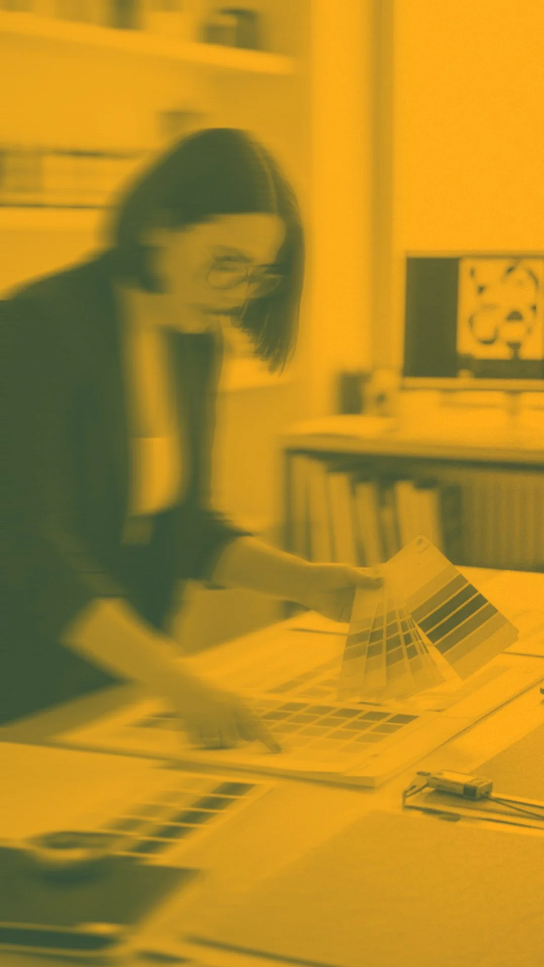 Une femme portant des lunettes en train de manipuler des échantillons de couleurs ou de peintures sur une table dans un espace intérieur, avec un ordinateur à écran plat en arrière-plan.