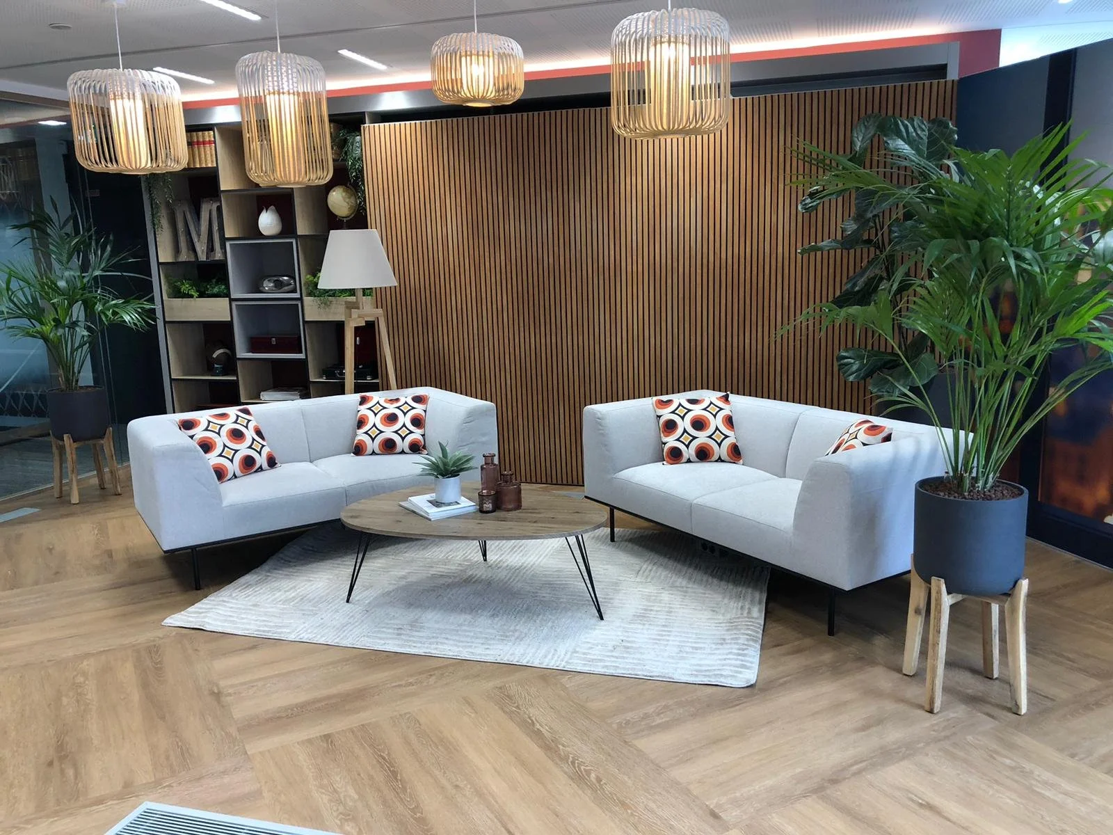Un salon moderne avec deux canapés blancs, une table basse en bois, plusieurs plantes vertes en pot, un tapis beige, et une décoration boisée verticale au mur.