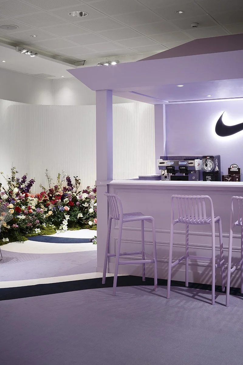 Un comptoir de café dans un espace moderne avec des chaises en plastique blanc, un mur avec un logo Nike en arrière-plan et un arrangement floral coloré.